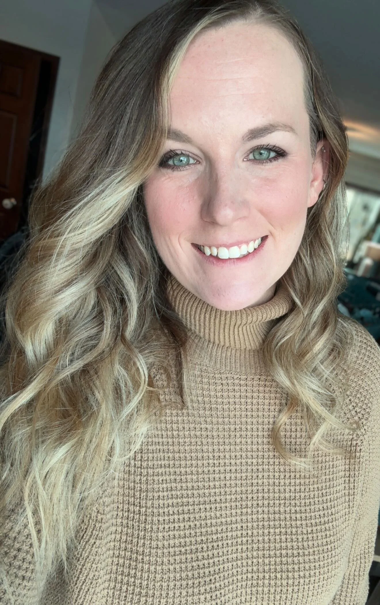 Close-up portrait of a smiling woman with light skin, blue eyes, long wavy blonde hair, wearing a beige turtleneck sweater, indoors with blurred background.
