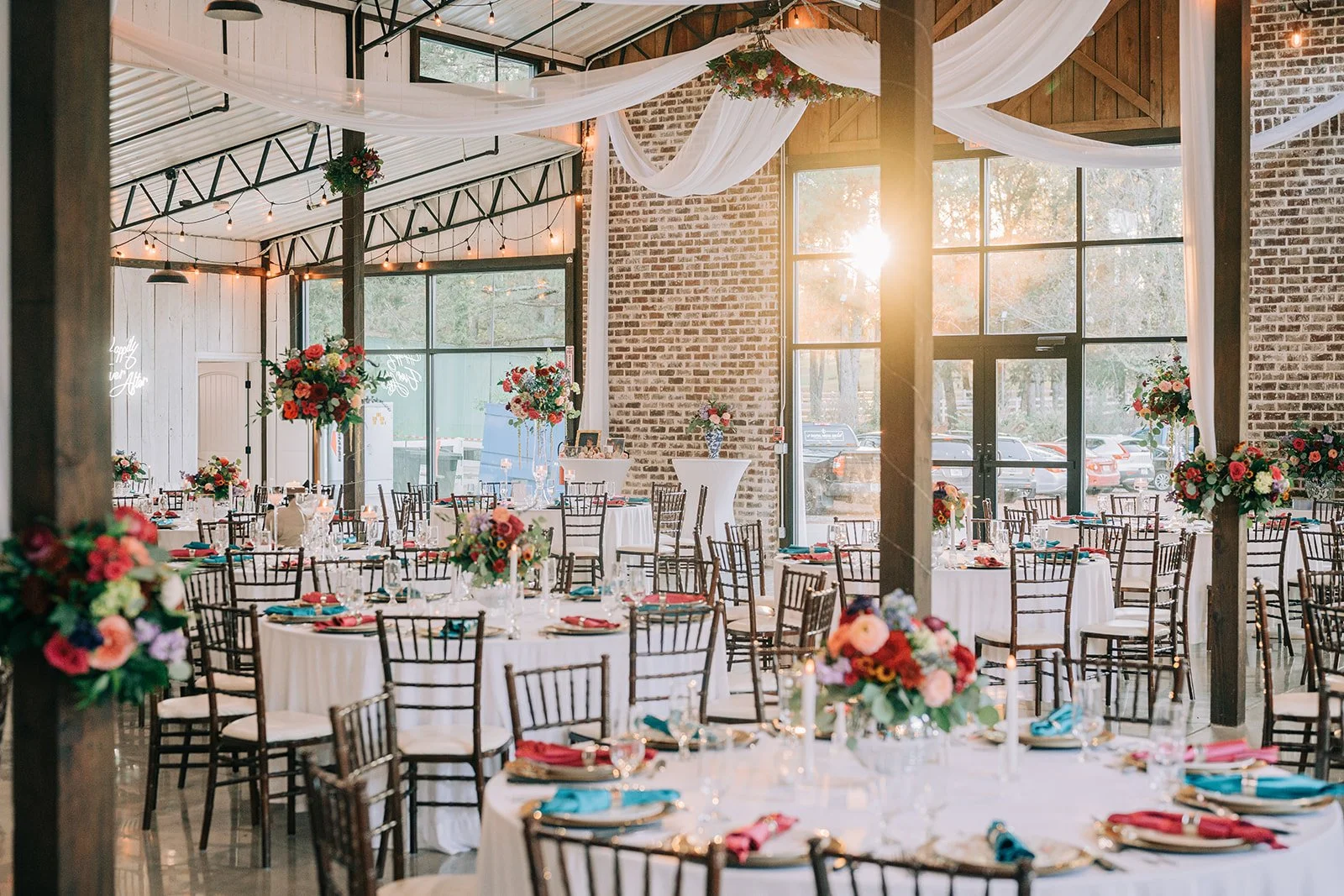 Wedding reception setup in a rustic barn with decorated tables, floral centerpieces, draped white fabric, exposed brick walls, large windows, and string lights.