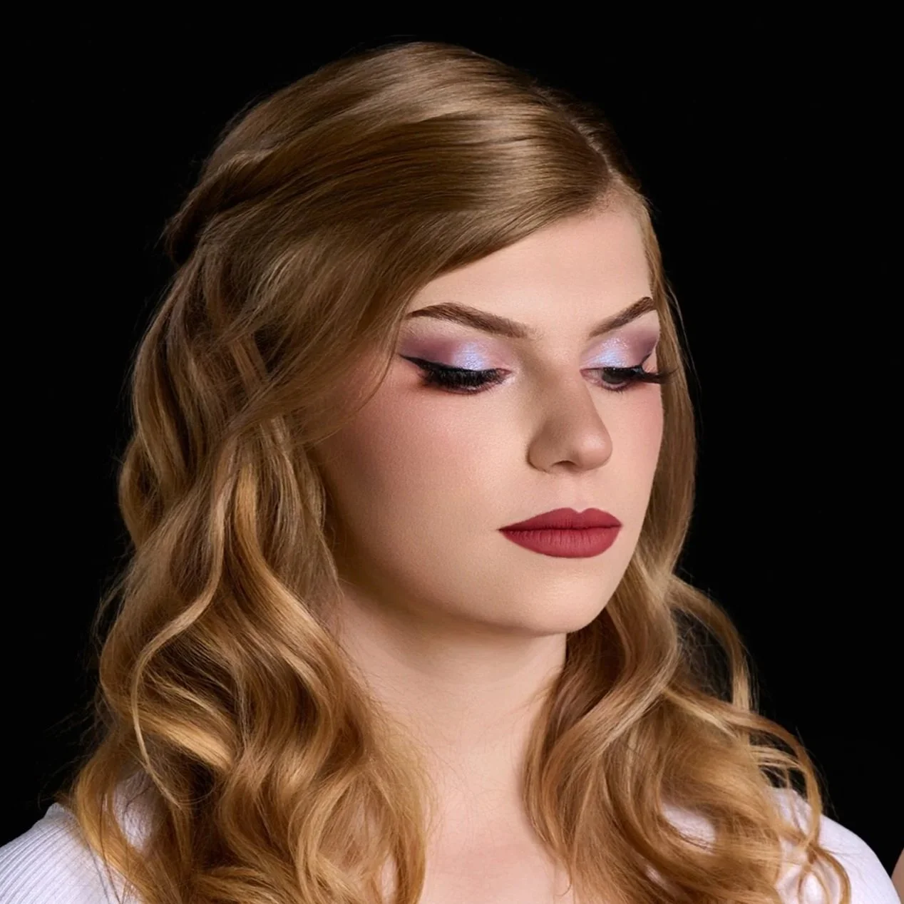 A young woman with long, wavy, light brown hair and makeup, including pink eyeshadow, black eyeliner, and red lipstick, against a black background.