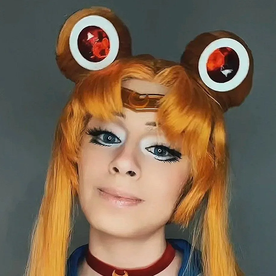 Person with long orange hair wearing a frog-themed headband with large eyes and a choker, making a playful facial expression.