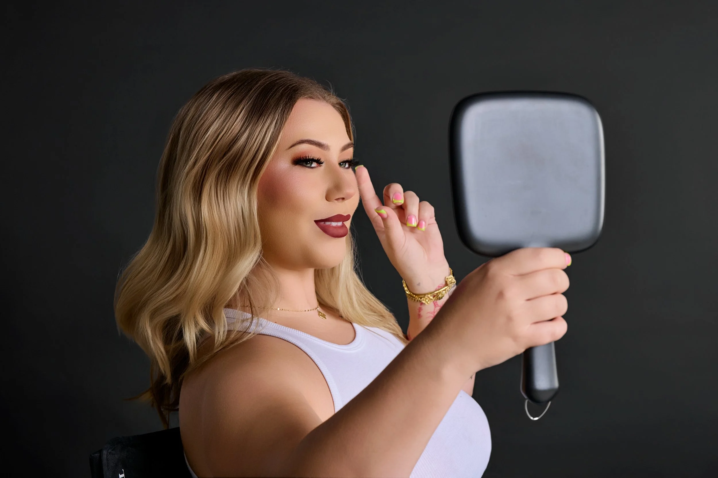 A woman with blonde hair, wearing a white tank top, applying makeup in front of a mirror with dark background.