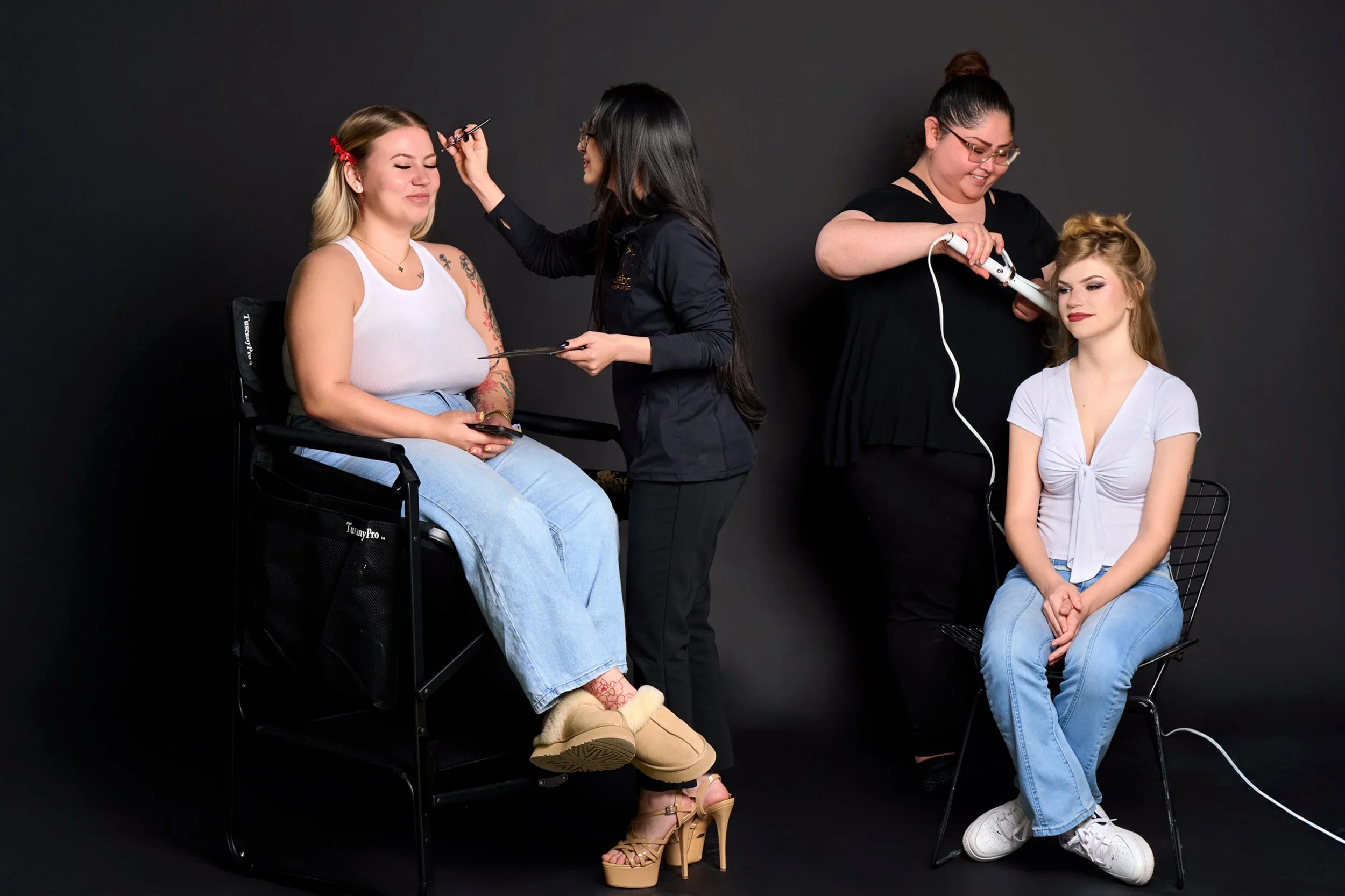 Two women are getting their makeup and hair done by two beauty artists in a studio with a black background. One woman is seated on a chair, with her eyes closed, while the other woman is seated on a wire chair, with her eyes open.