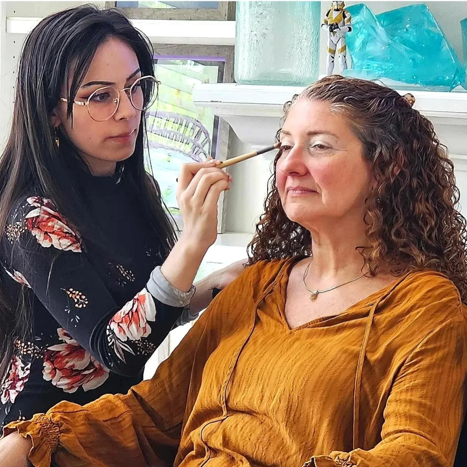 A woman with curly brown hair and a gold necklace sitting comfortably while makeup artist applies makeup near her eye, near a white mantel with blue and green decorations in the background.