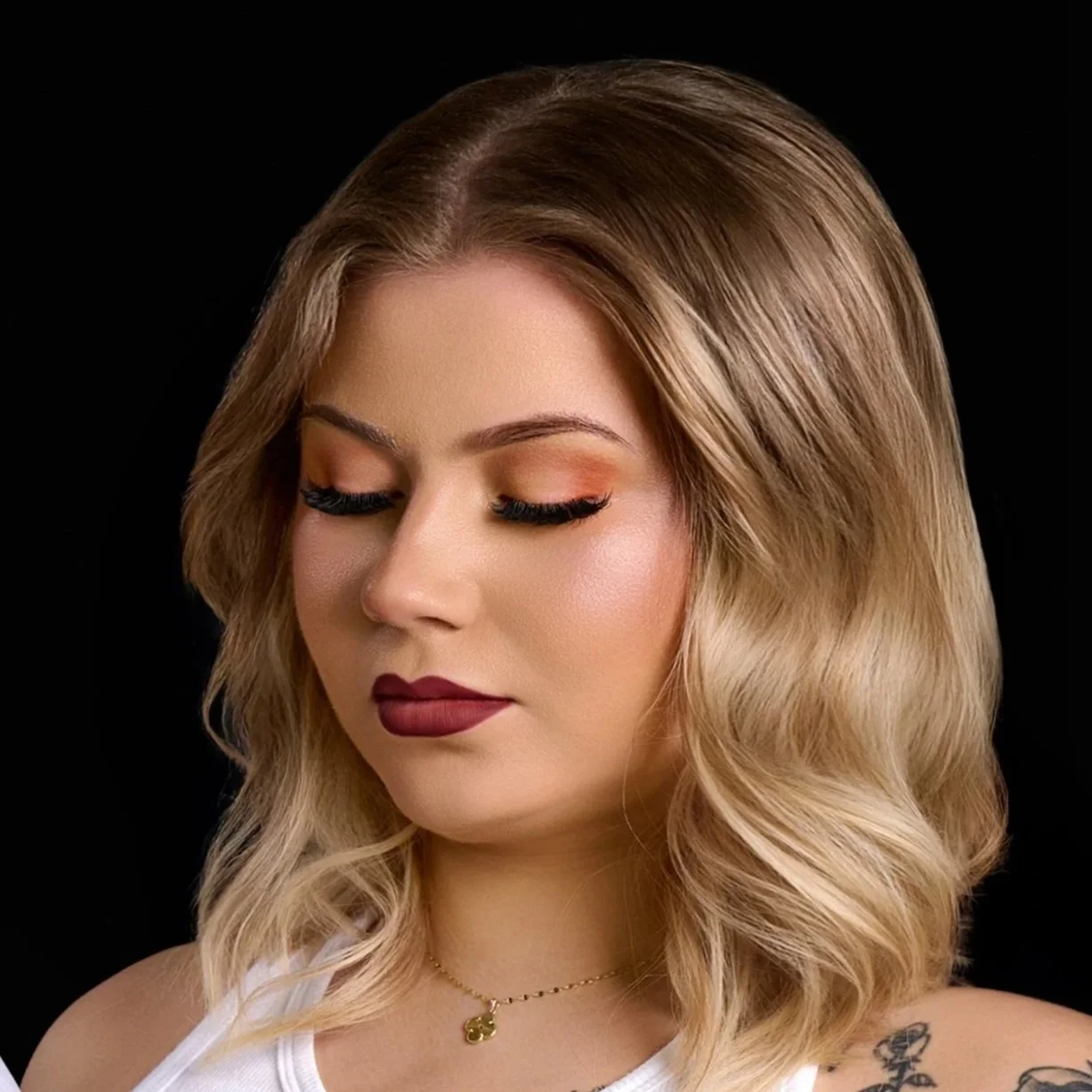 A woman with shoulder-length wavy blonde hair, closed eyes, makeup, and dark lipstick, wearing a white tank top and a gold necklace with a small pendant, posing against a black background.