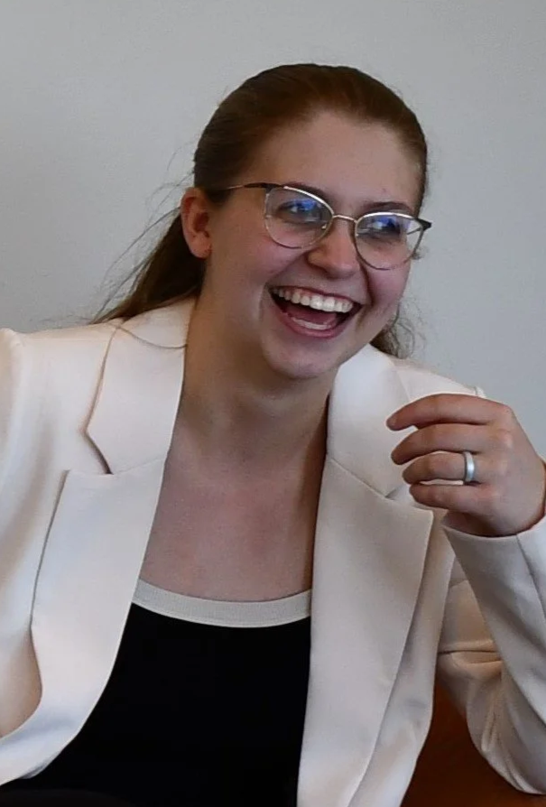 A woman with glasses and a white blazer laughing and smiling, seated indoors.