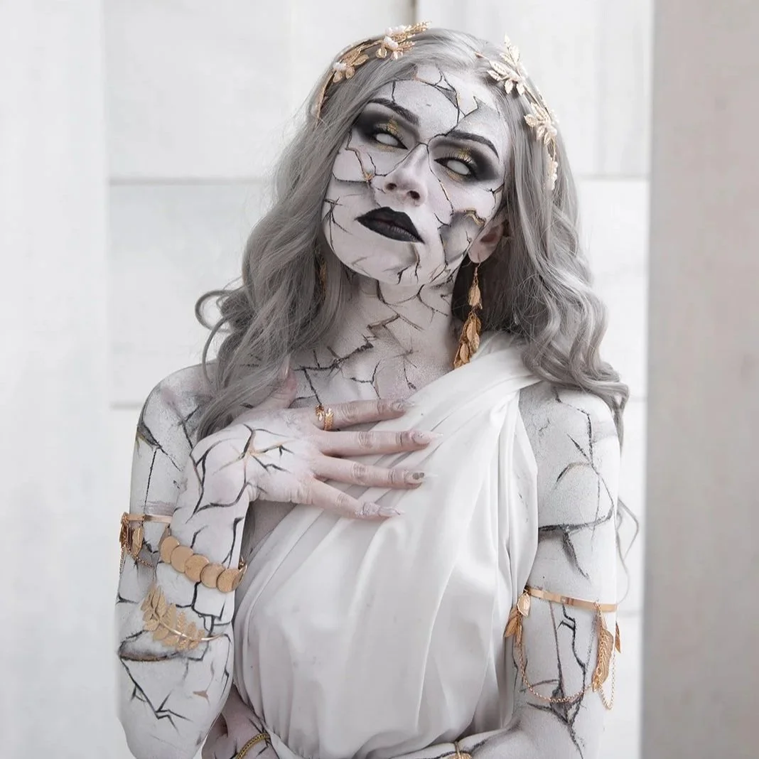 Person with cracked white face makeup, gray hair, black lipstick, gold jewelry, dressed in white, posing against a light background.