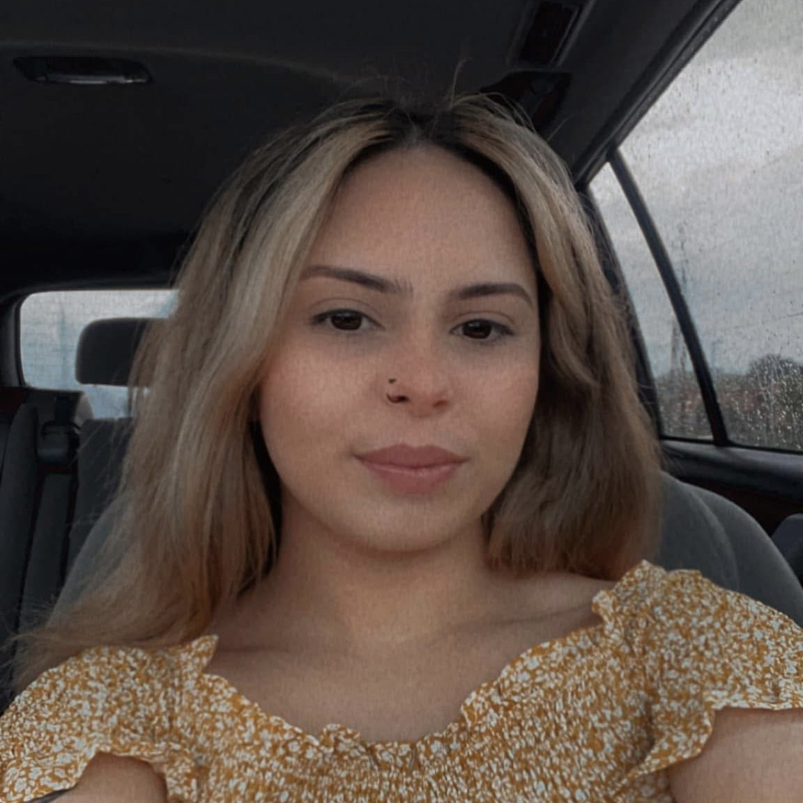 A woman with light brown hair and a small nose piercing sitting in a car with rain on the window behind her, wearing a yellow floral off-shoulder top.