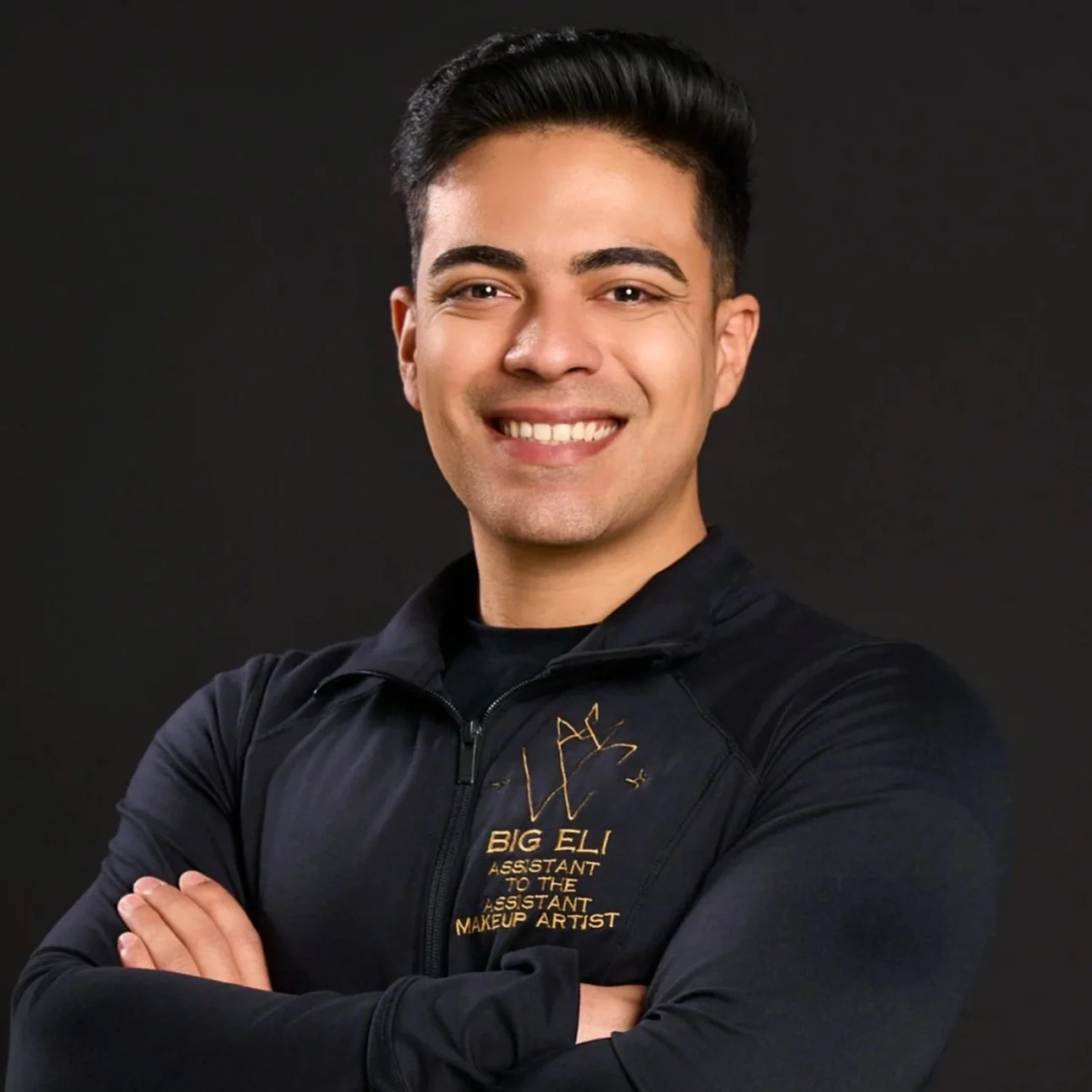 Smiling man with crossed arms wearing a black jacket with gold embroidery and logo, standing against a dark background.