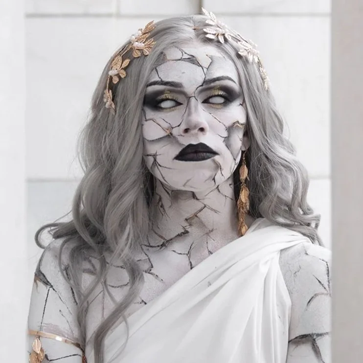 Person with cracked white and black face makeup, gold floral headpiece, white toga, and long earrings, resembling a marble statue or cracked stone figure.