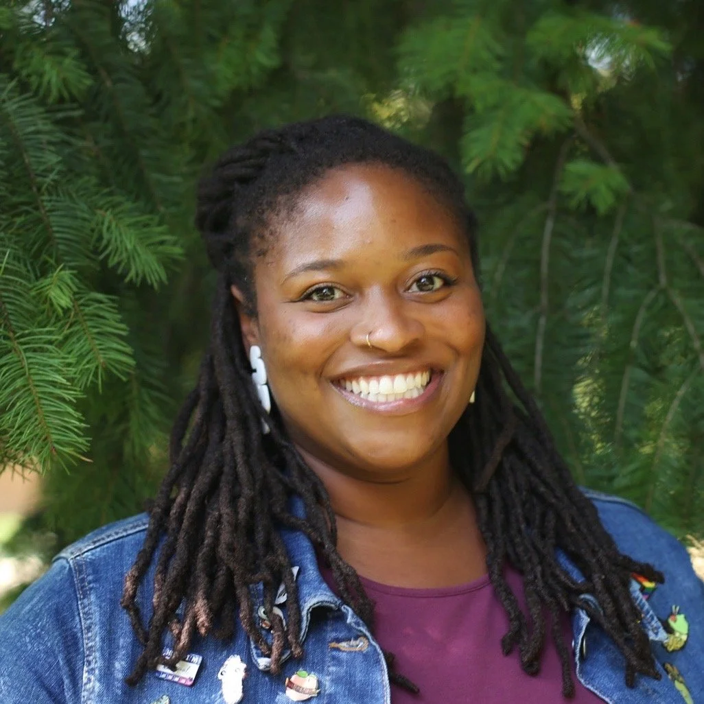 A woman with dark skin and dreadlocks smiling outdoors in front of green tree branches.