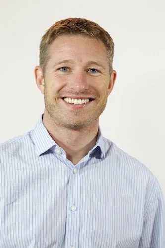 A man with short light brown hair, blue eyes, and a light complexion smiling. He is wearing a light blue collared shirt and standing against a plain white background.