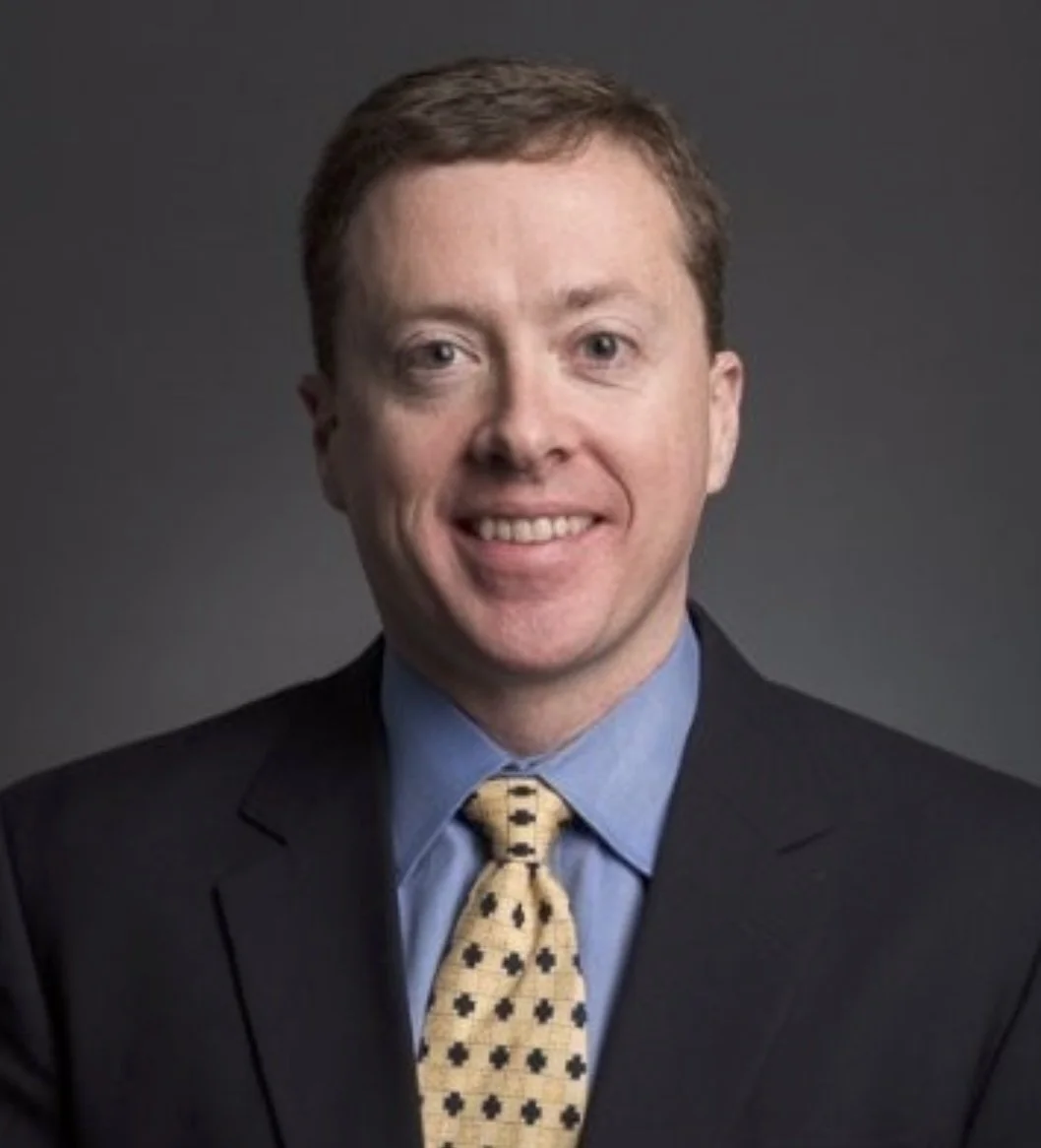 Portrait of a man with short brown hair, wearing a dark suit, light blue shirt, and a yellow tie with black diamond shapes, smiling at the camera against a dark gray background.