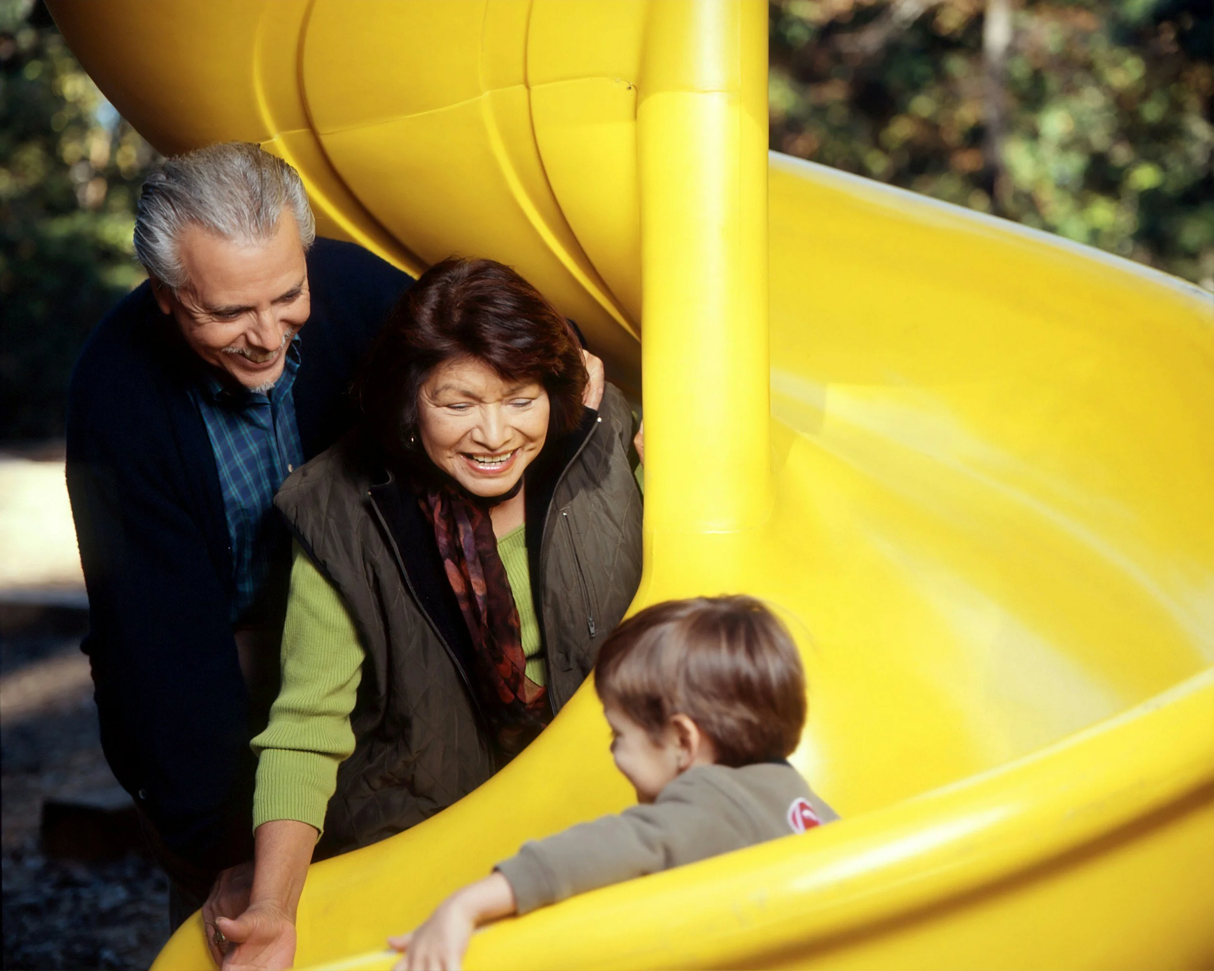 Two adults and a child are playing on a yellow slide in a park, with trees in the background.