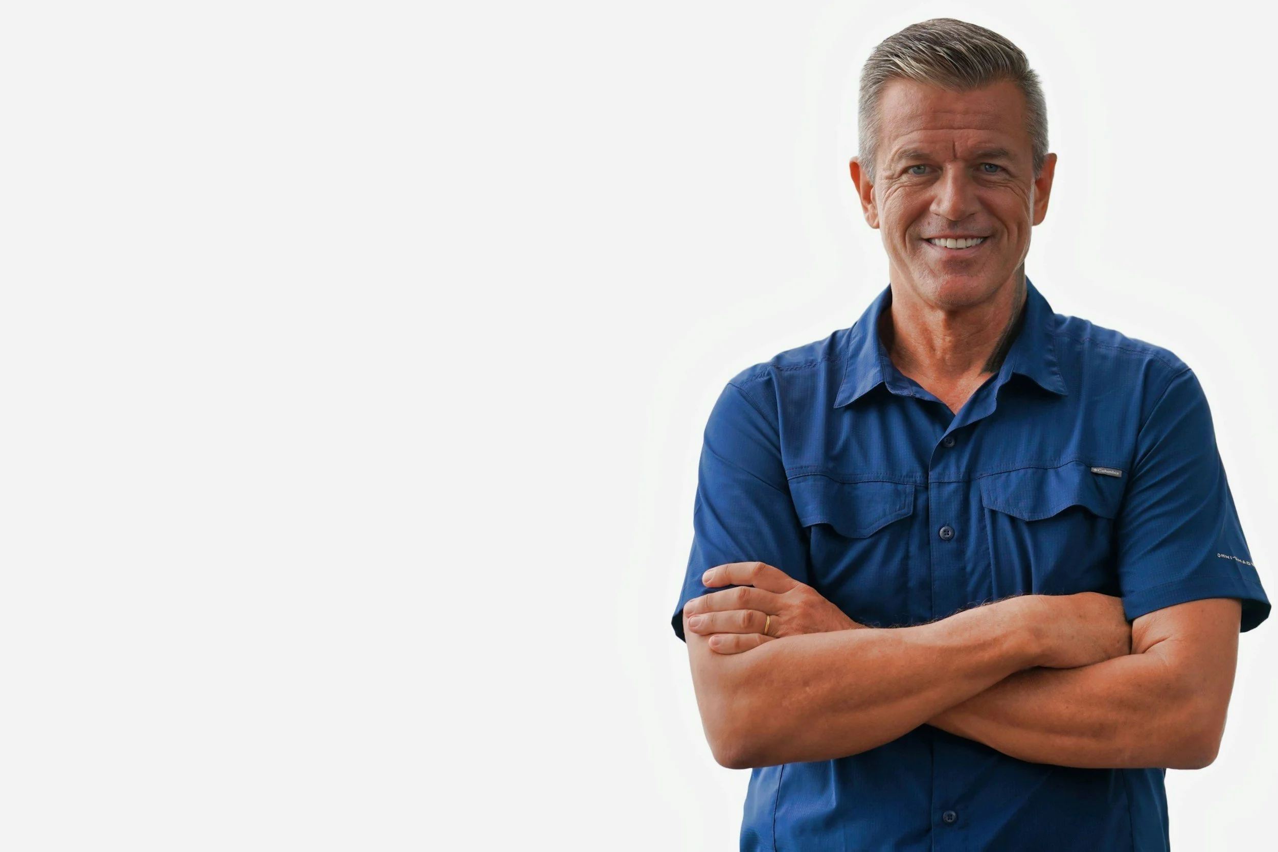 Smiling middle-aged man with short gray hair, wearing a blue button-up shirt with rolled-up sleeves, standing with arms crossed against a white background.