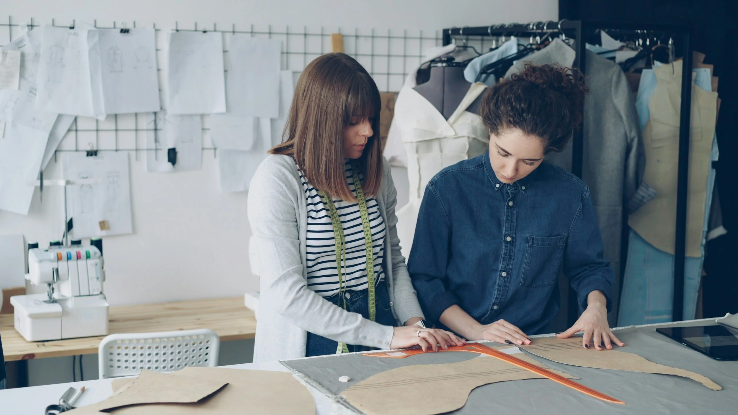Two women working together on a sewing project in a fashion design studio, with fabric, sewing machine, and sketches visible.