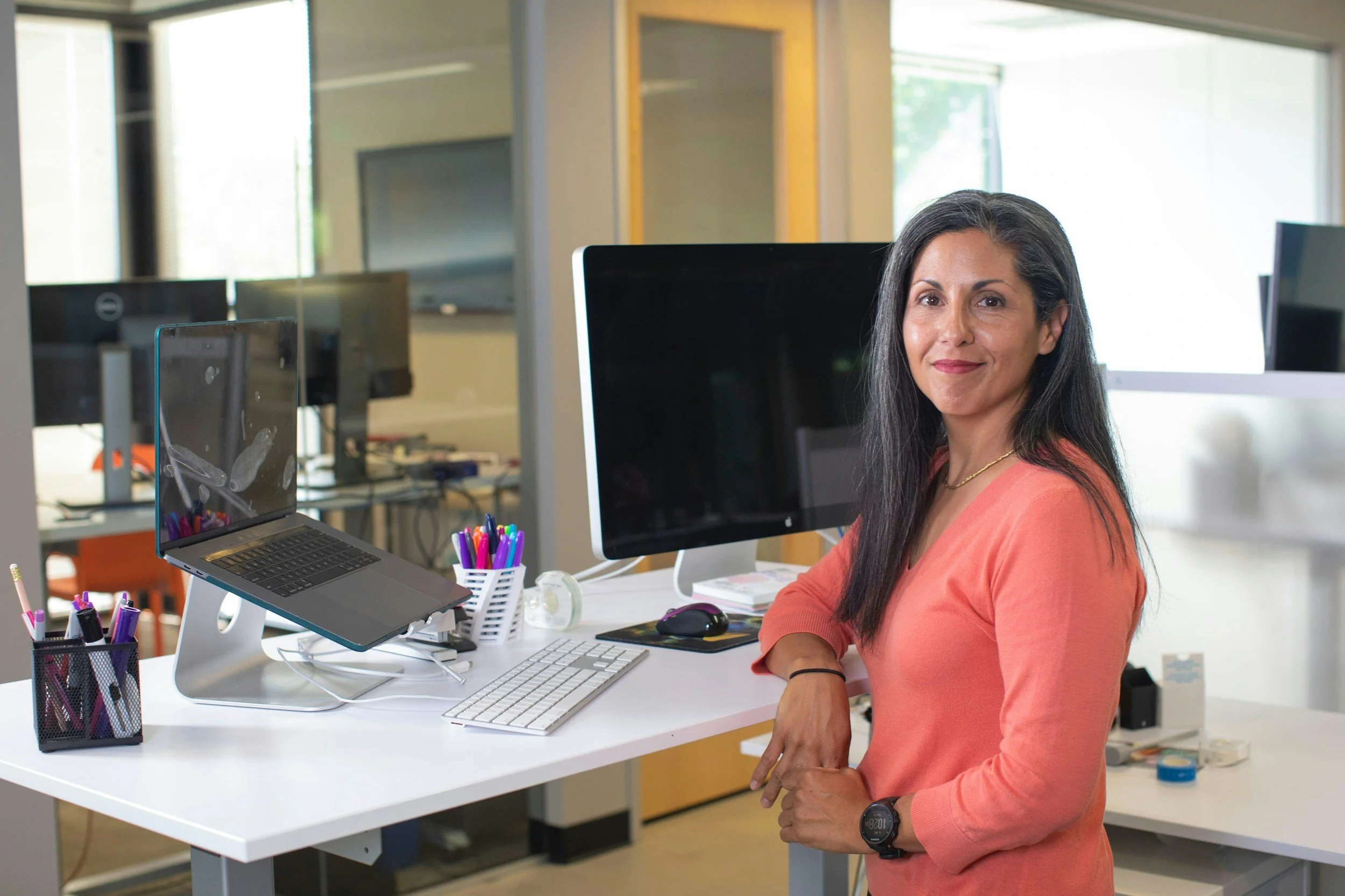A woman with long black hair wearing a pink sweater standing at a white desk in an office, smiling.