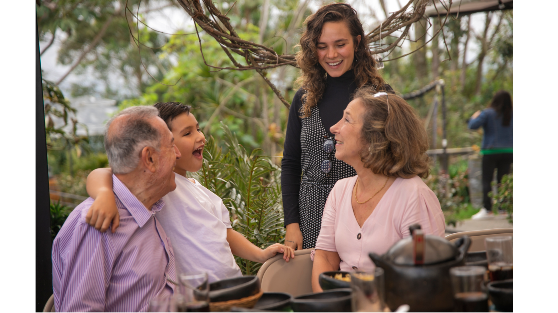 Family of four at a dining table outdoors, smiling and talking, surrounded by greenery and trees.