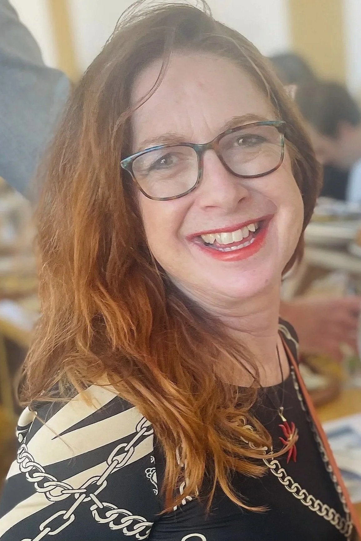 A woman with red hair, glasses, and a bright smile, wearing a patterned top with chain designs, standing indoors at a social event.