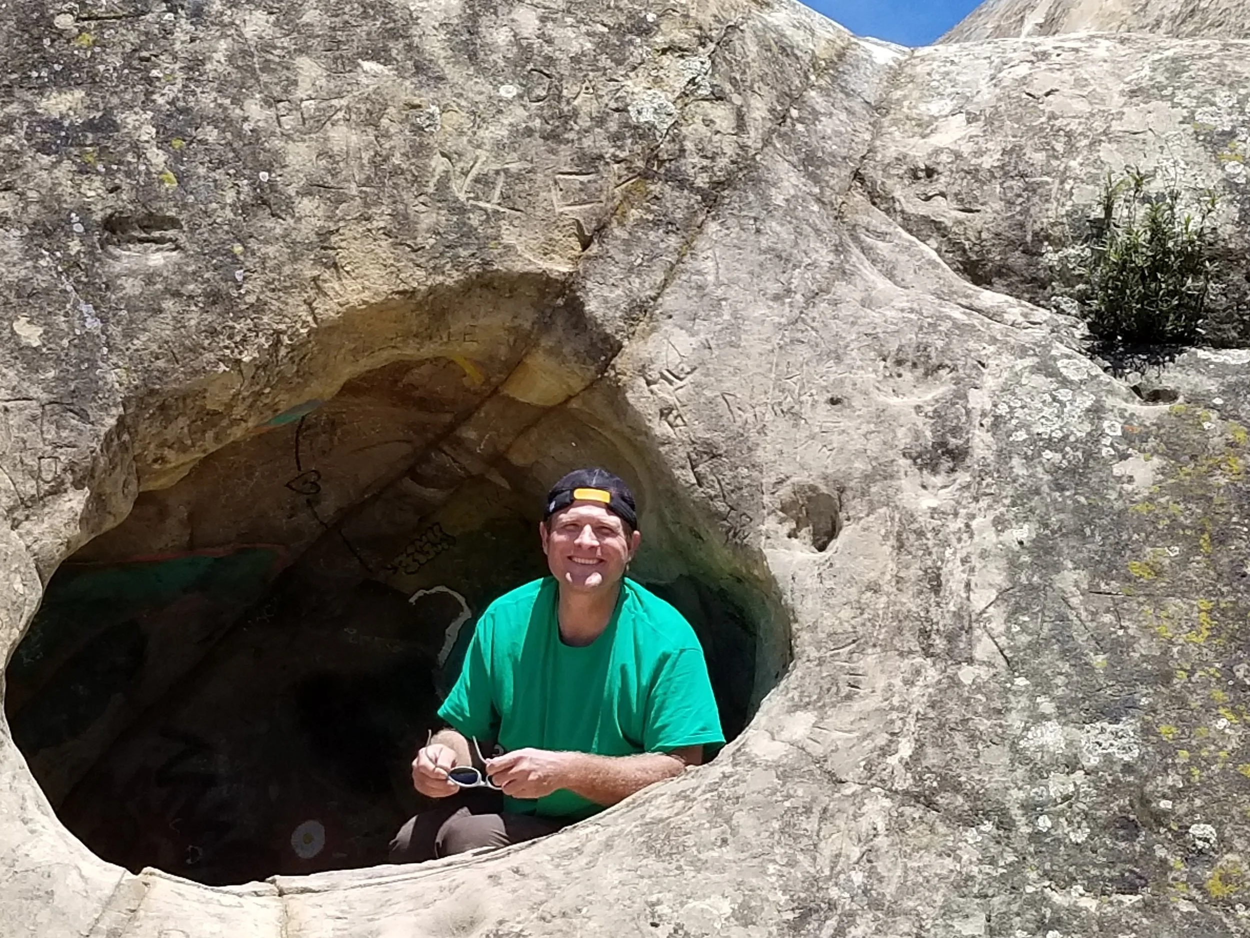A man wearing a green shirt and a black cap kneels inside a large rock formation with a natural circular hole, smiling at the camera, with a clear blue sky overhead