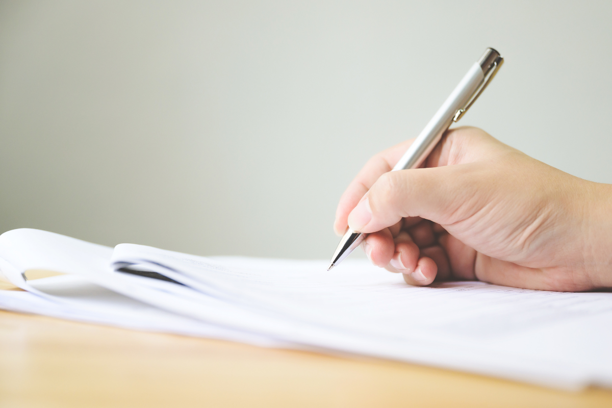A person's hand holding a silver pen, writing on white paper with multiple sheets, on a wooden surface.