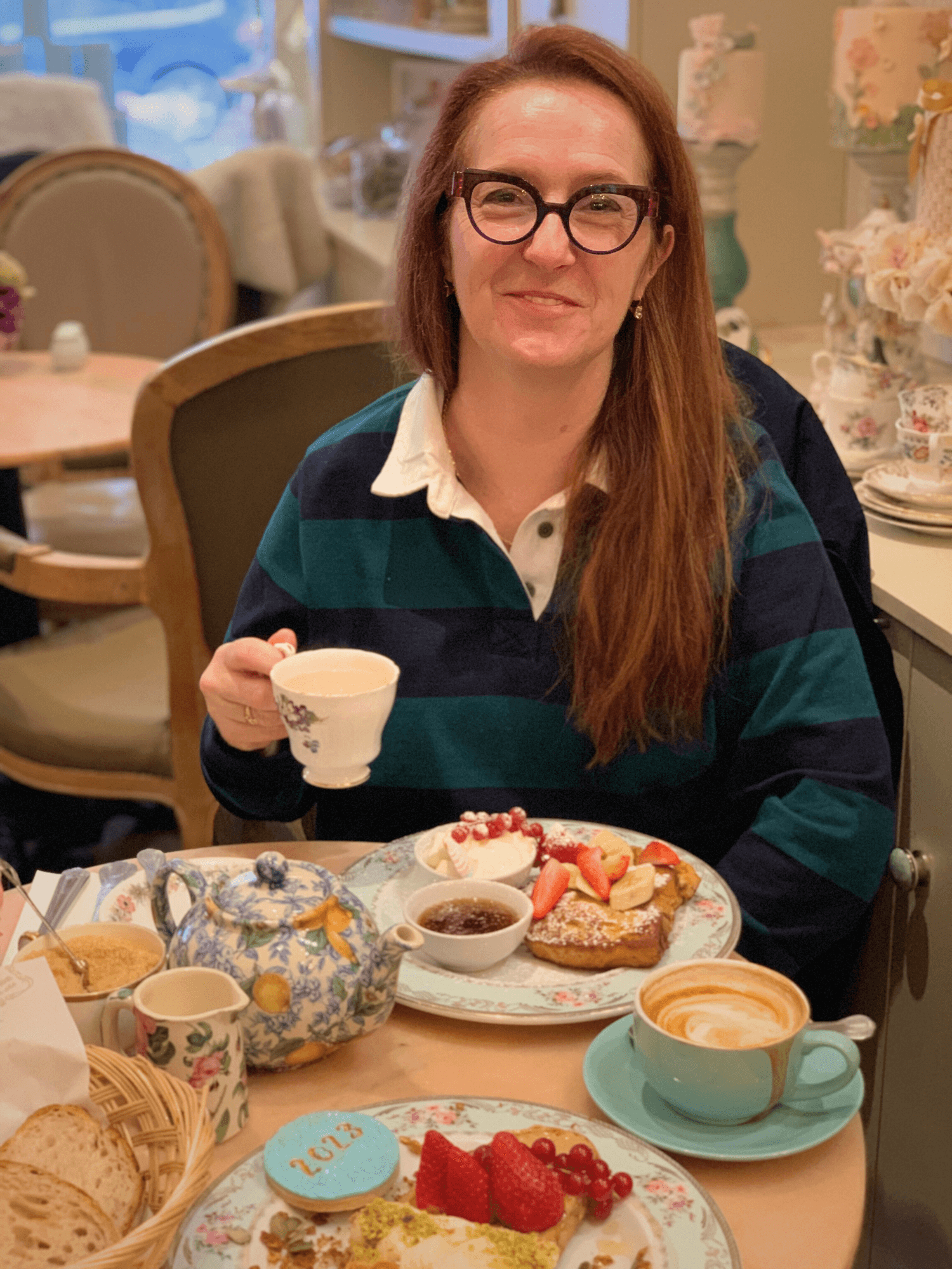 A woman with glasses and long red hair sitting at a table with a tea set, coffee, and desserts, holding a teacup and smiling in a cozy café setting.