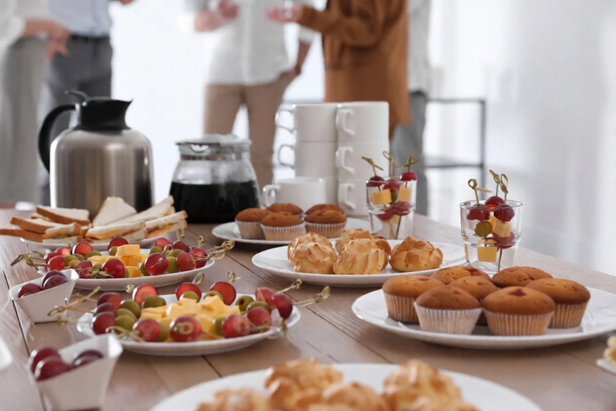 A table set with assorted breakfast foods including muffins, cupcakes, fruit skewers, and beverages, with people in the background.