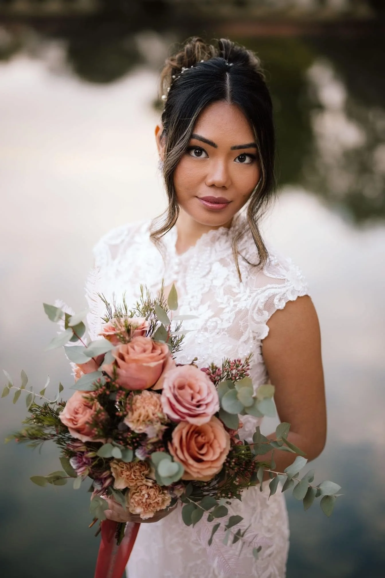 A bride holding a bouquet of pink roses and greenery outdoors near a body of water at sunset.