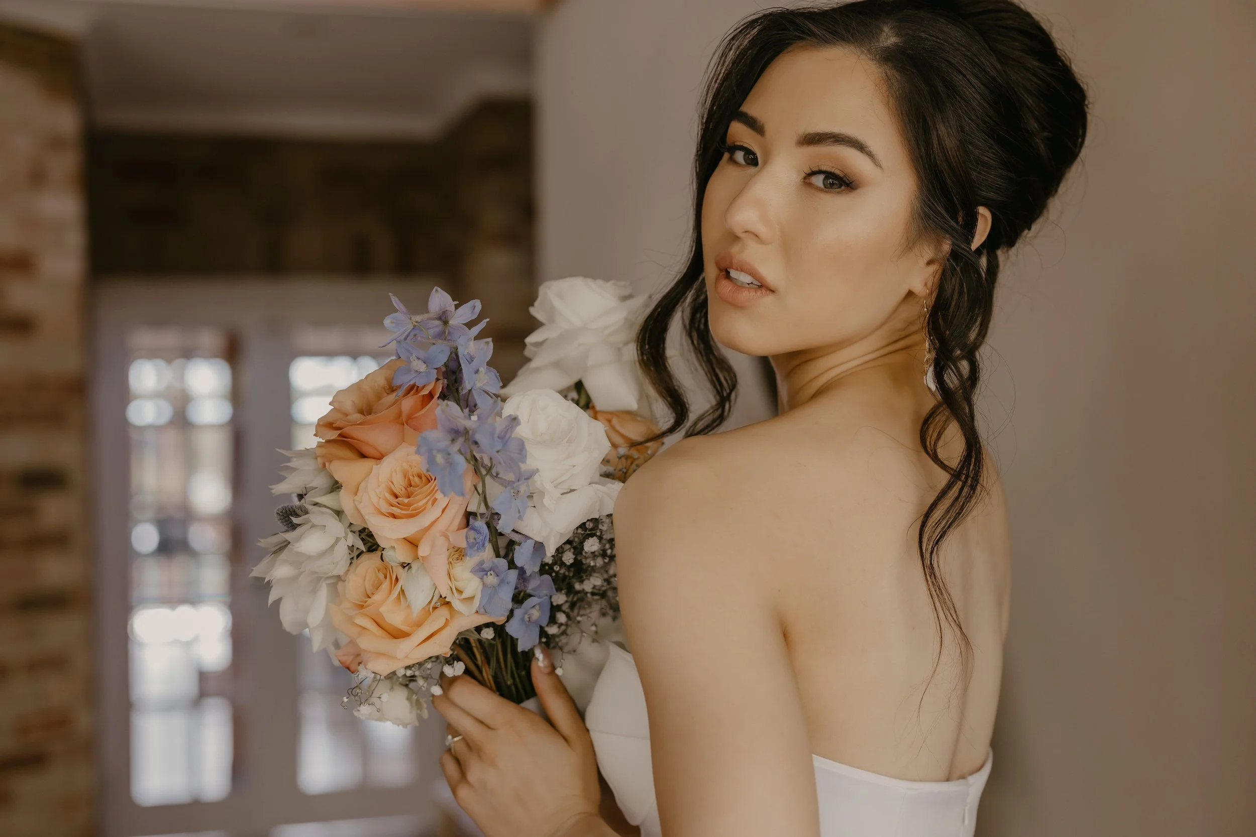 A young woman with dark hair styled in loose waves holds a bouquet of pastel-colored flowers, including peach roses, white flowers, and purple blooms, while looking over her shoulder at the camera inside a room.
