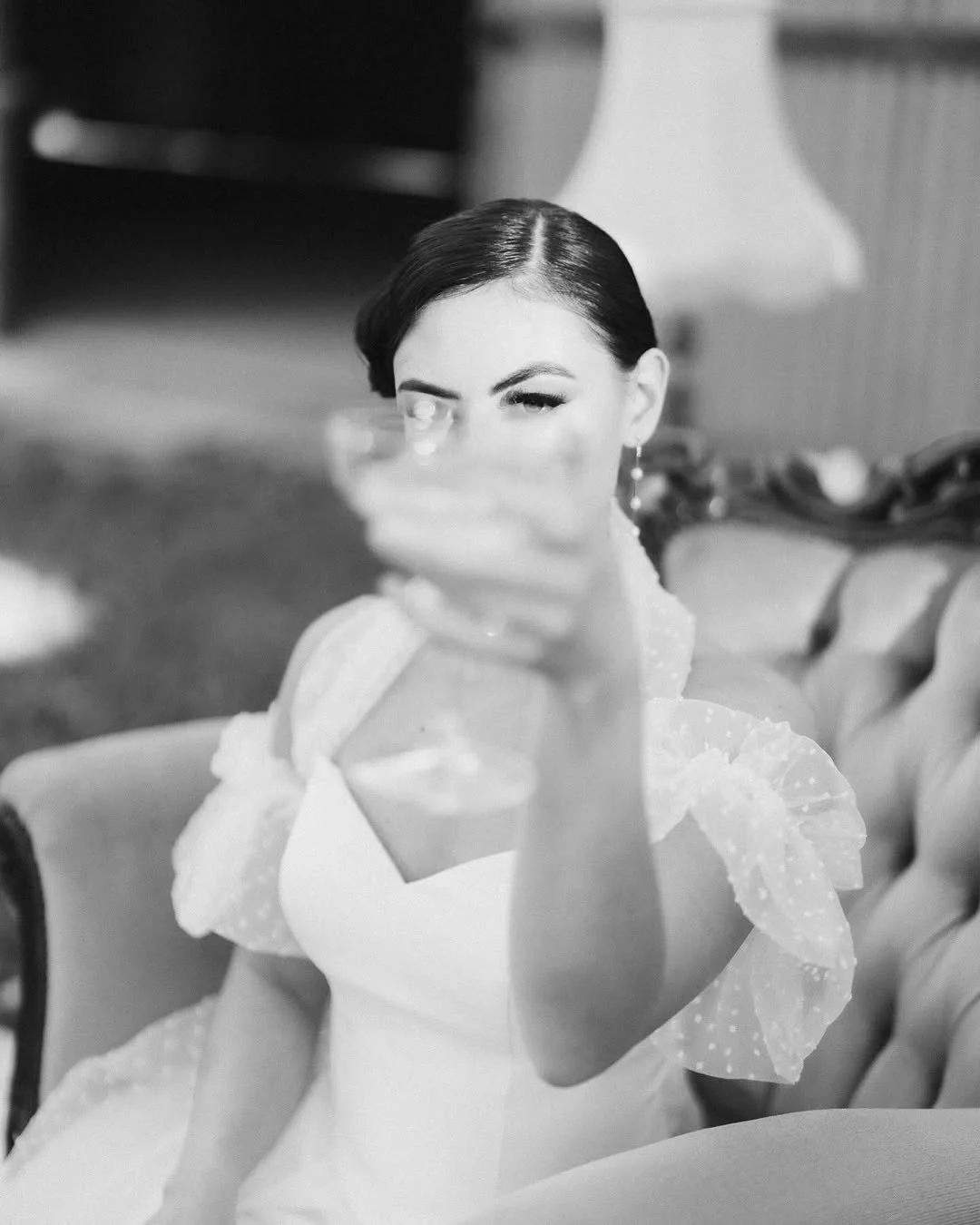 A woman with dark hair and makeup holding a glass of drink, sitting on a vintage-style sofa in a room with a lamp in the background.