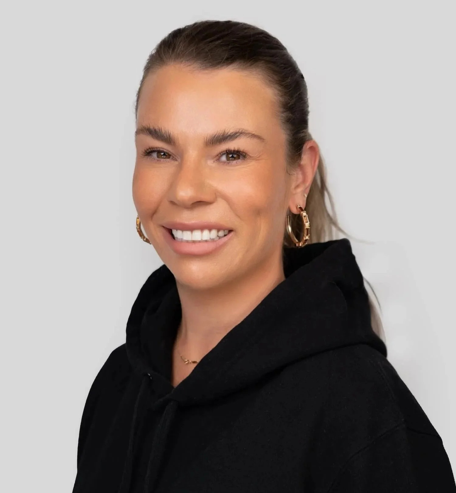 A smiling woman with brown hair tied back, wearing hoop earrings and a black hoodie with a logo that says 'TALS' on it, against a light gray background.