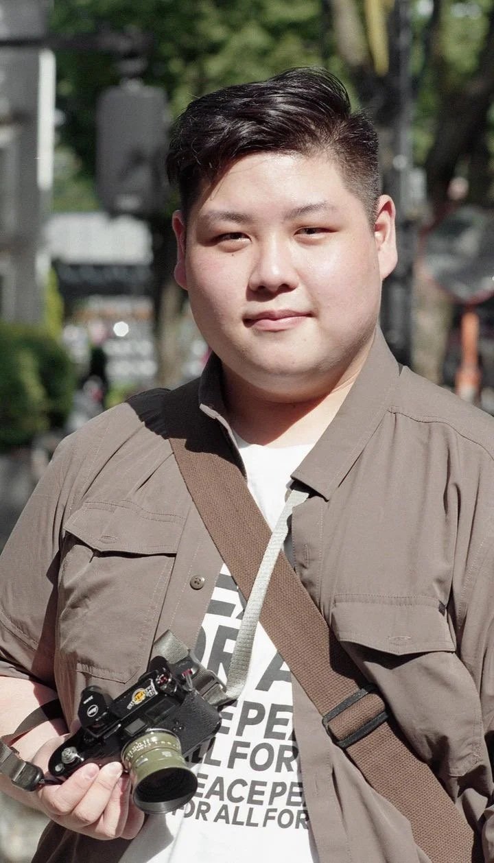 A young man holding a camera outdoors with trees and blurred background