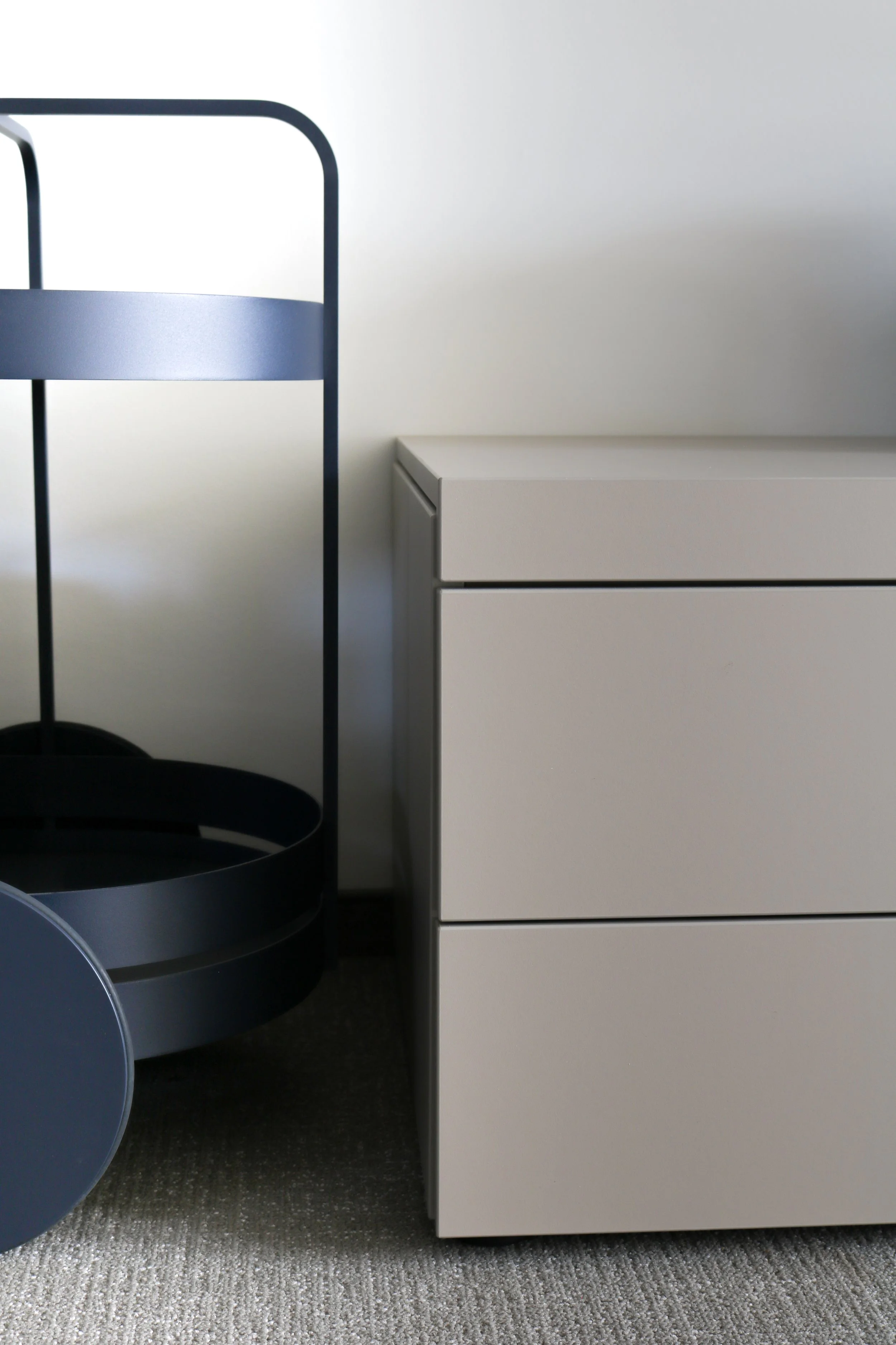 Ein weißer Schrank neben einem runden, schwarzen Servierwagen auf einem grauen Teppichboden.