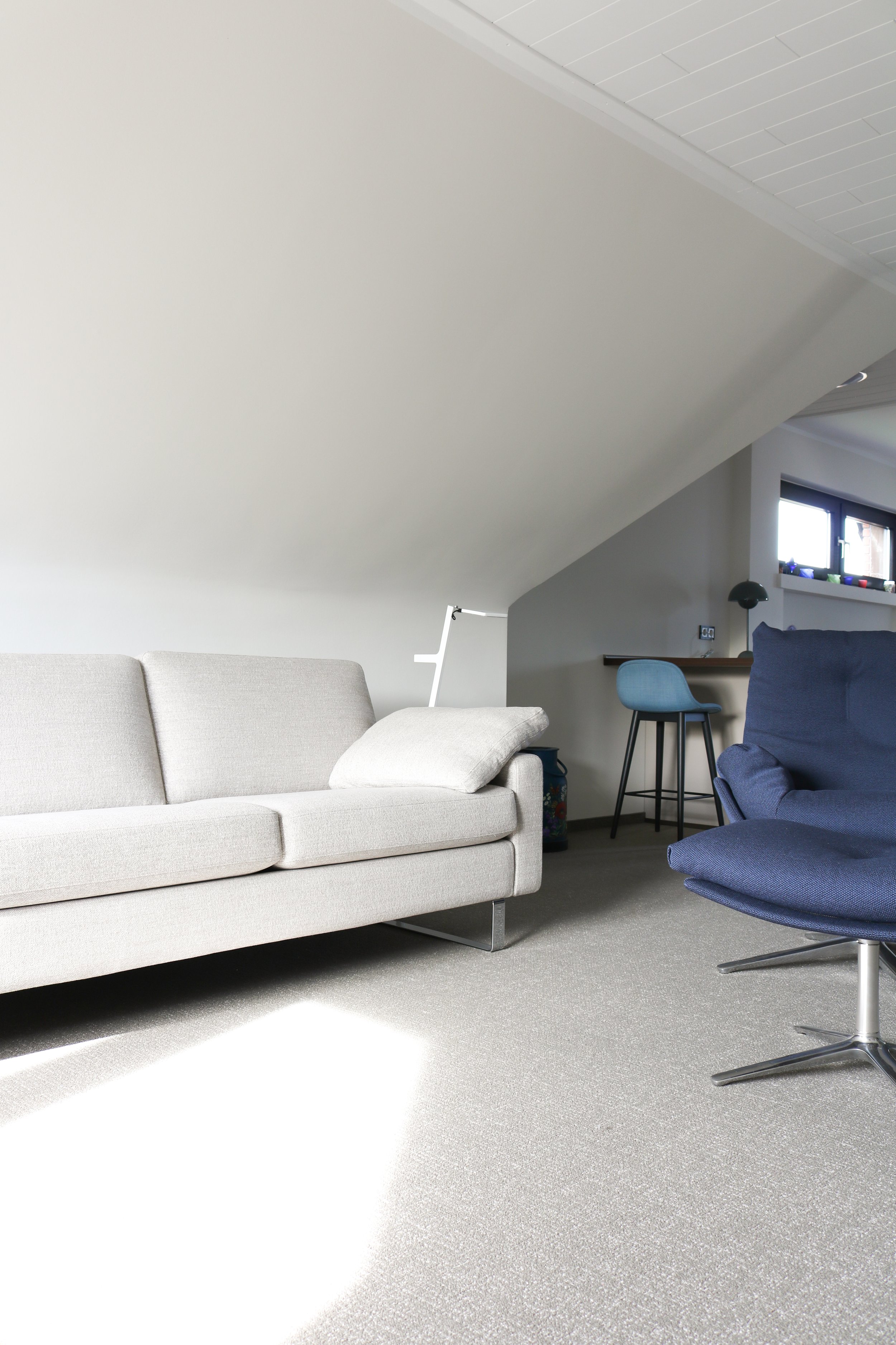 Ein Wohnzimmer mit grauem Sofa, blauer Ledercouch, Stehlampe, Trittstufe und einem kleinen Arbeitsbereich mit Stuhl und Tisch unter einem Dachfenster.