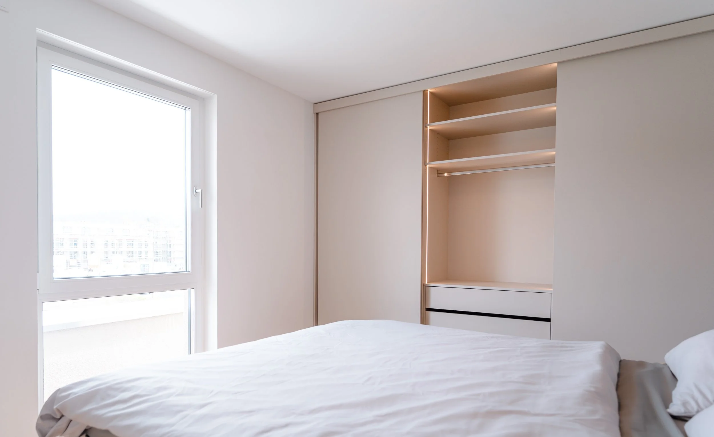 Ein modernes, minimalistisch gestaltetes Schlafzimmer mit weißem Bett, einem großen Fenster und einem eingebauten Kleiderschrank mit offener Regal- und Schrankfläche.
