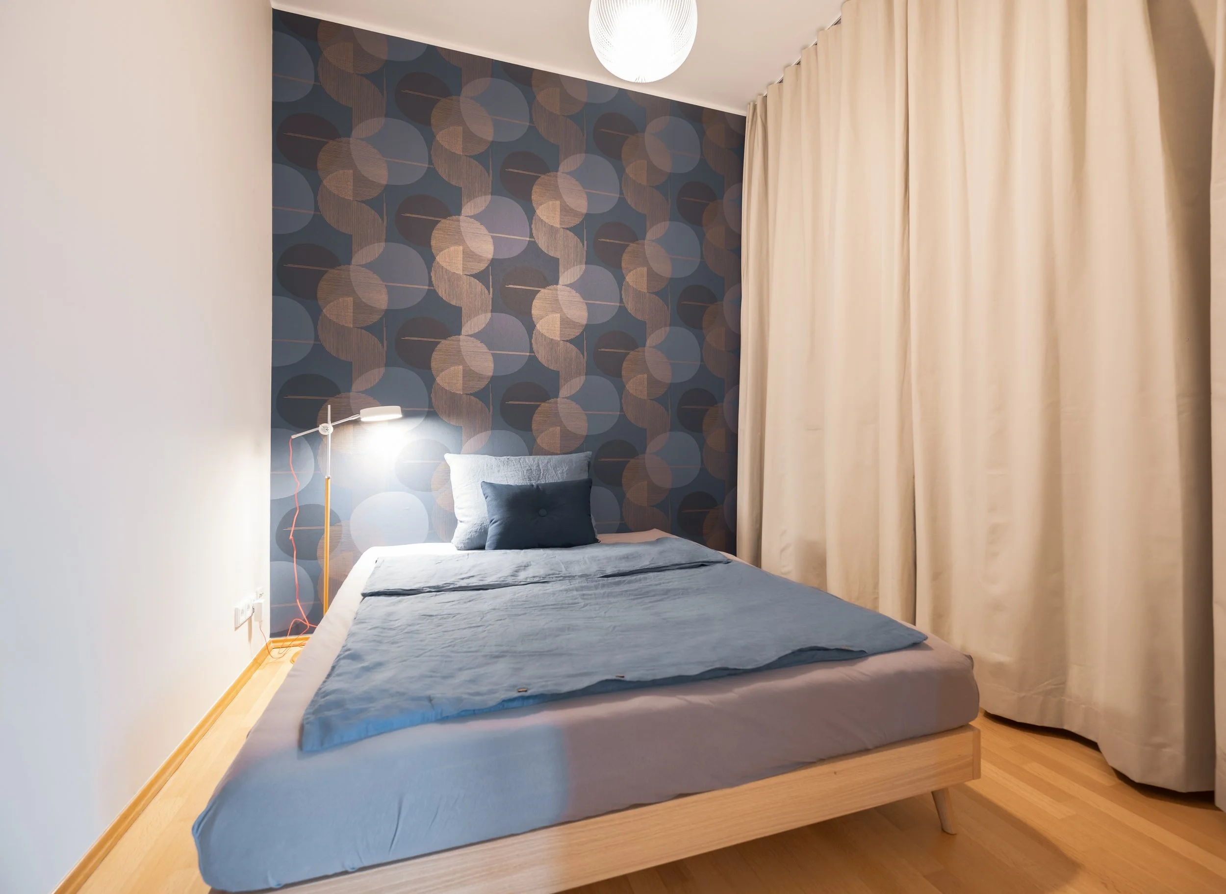 Schlafzimmer mit blaue Bettwäsche, einer blau-gestreiften Kissen, einer Lampe links und hellen Vorhängen an der rechten Seite, Wand mit geometrischem Muster.
