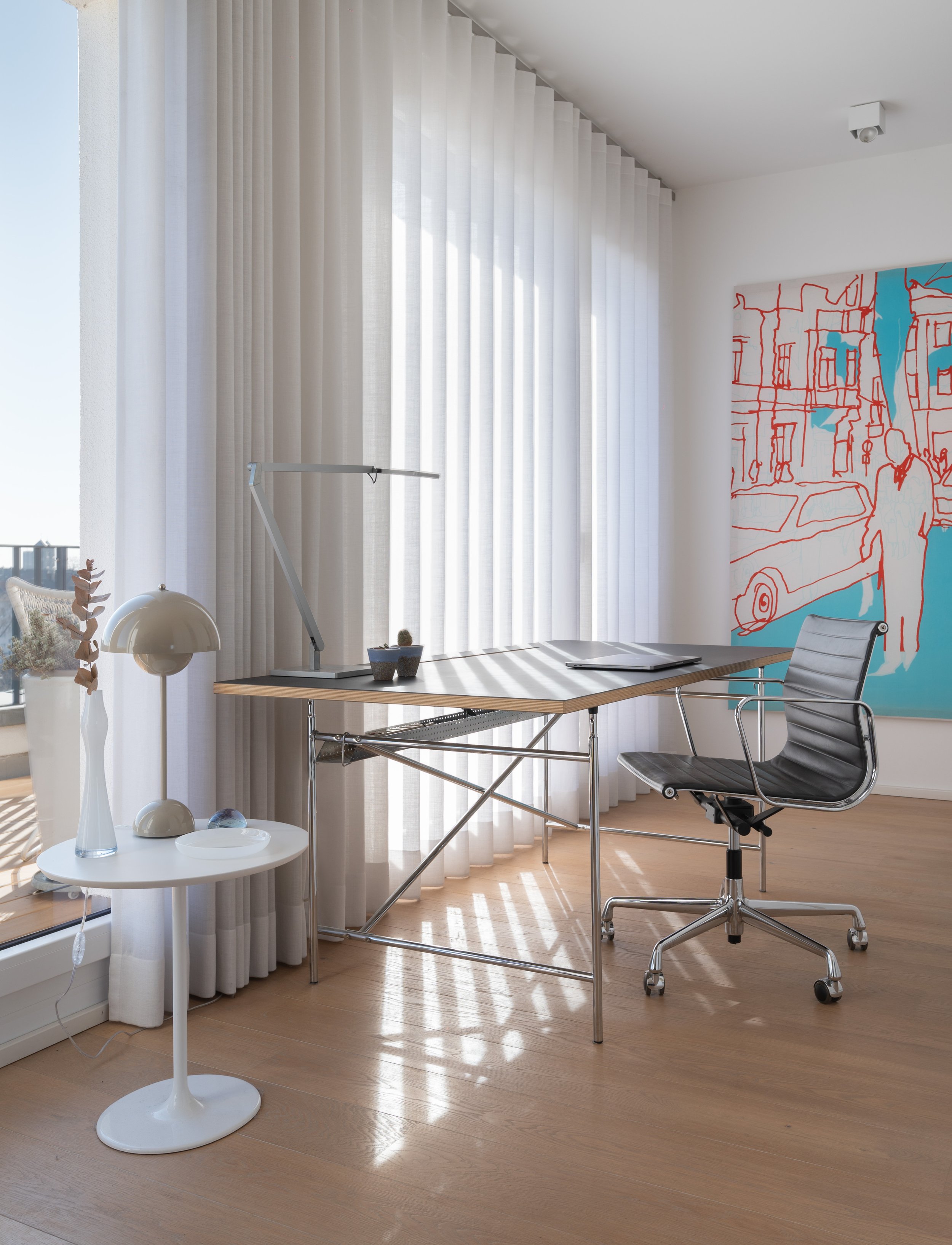 Schreibtisch in einem hellen, modernen Büro mit schwarzen Bürostuhl, Stehlampe, Kunstwerk an der Wand und weißen Vorhängen.