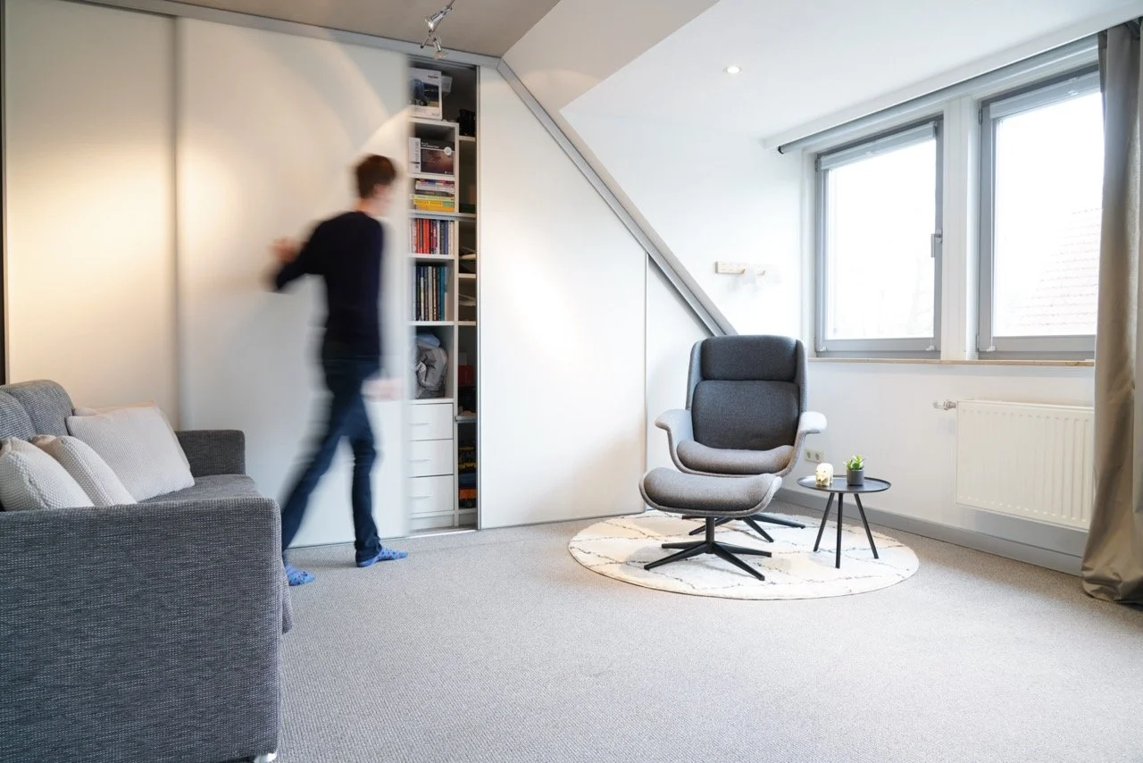 Modernes Wohnzimmer mit grauem Sessel, kleinen schwarzen Tischchen, Schreiner Einbauschrank Bücherregal, Couch und großen Fenstern