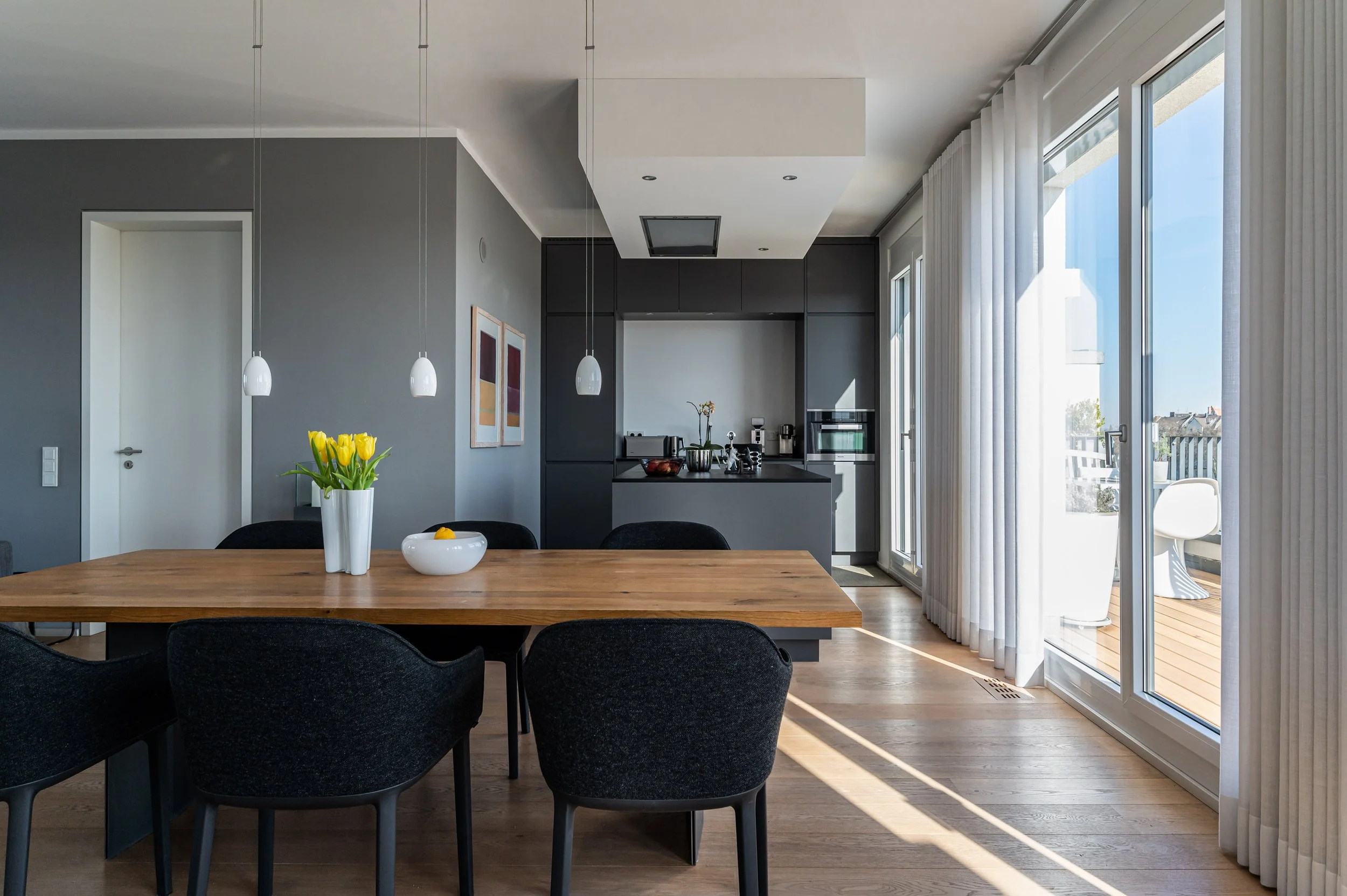 Moderne offene Küche mit Essbereich, Holztisch, schwarze Stühle, graue Wände, große Fenster mit weißen Vorhängen, Balkon sichtbar draußen.