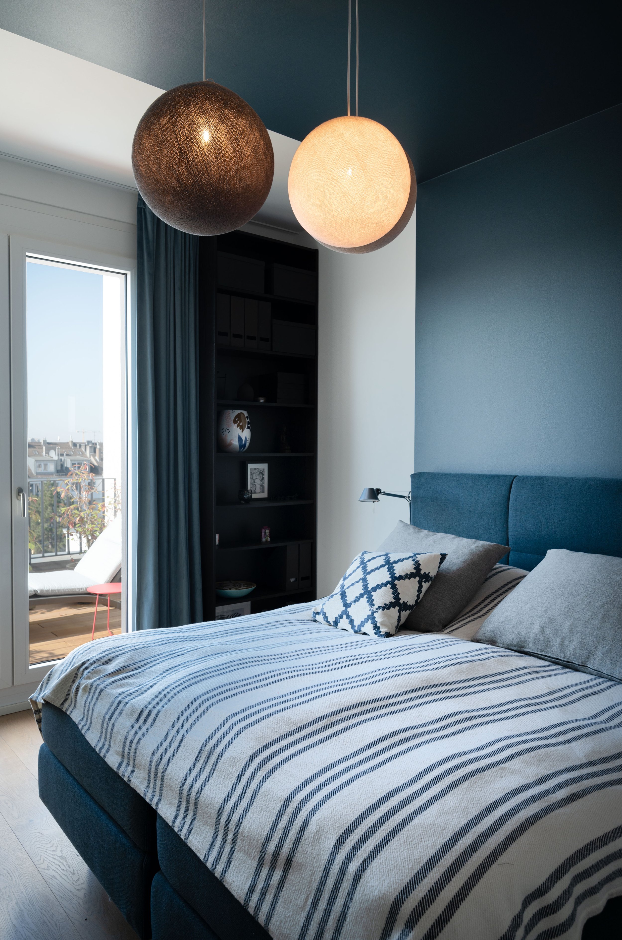 Schlafzimmer mit blauem Kopfteil, gestreiftem Bettzeug, Kissen, Gardinen, Regal und Balkon mit Sitzmöglichkeit