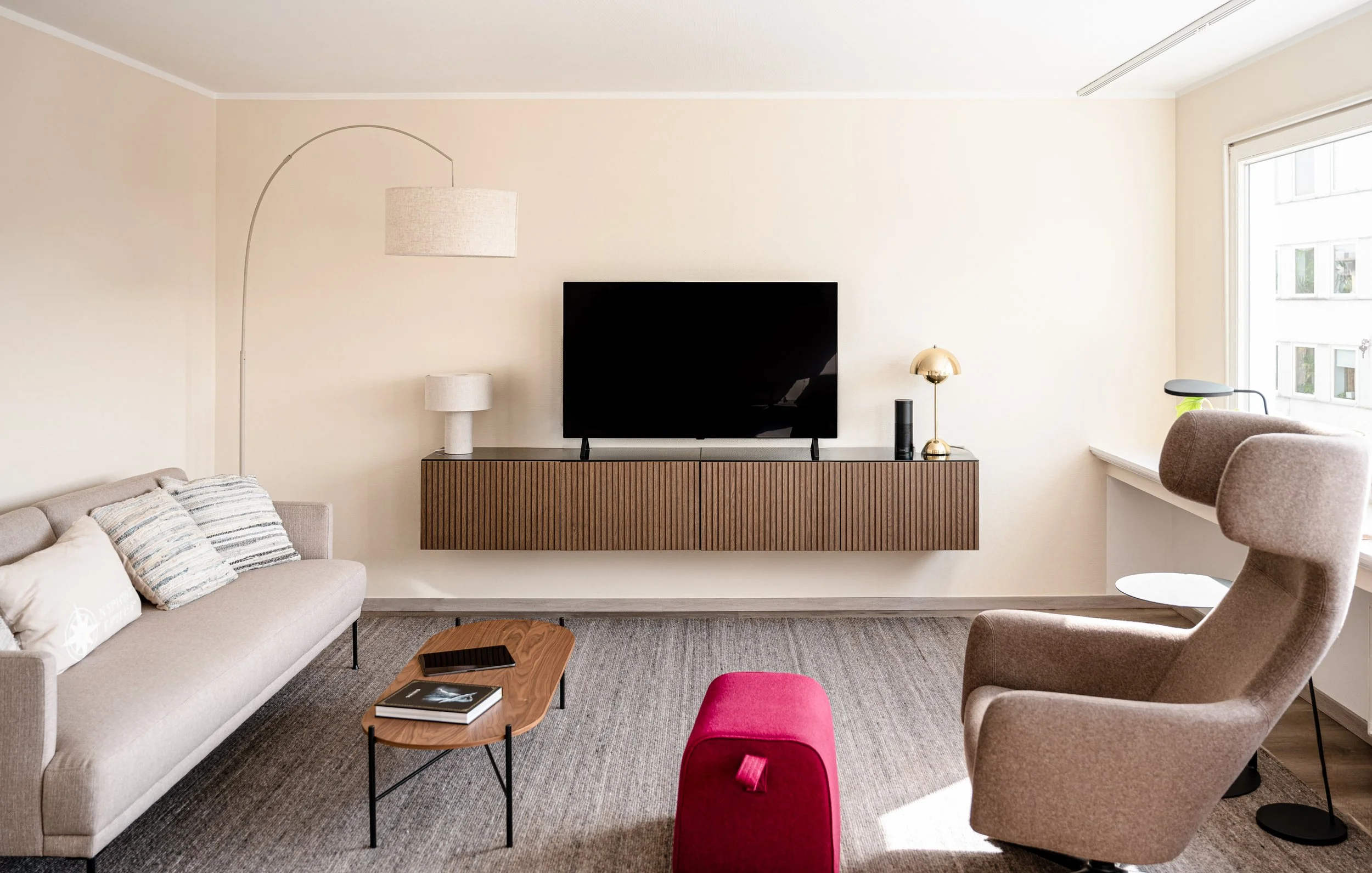 Moderner Wohnzimmerbereich mit braunem Sofa, buntem Hocker, Couchtisch, Fernseher an Wand, Schreibtischlampe, Stehlampe und großem Fenster