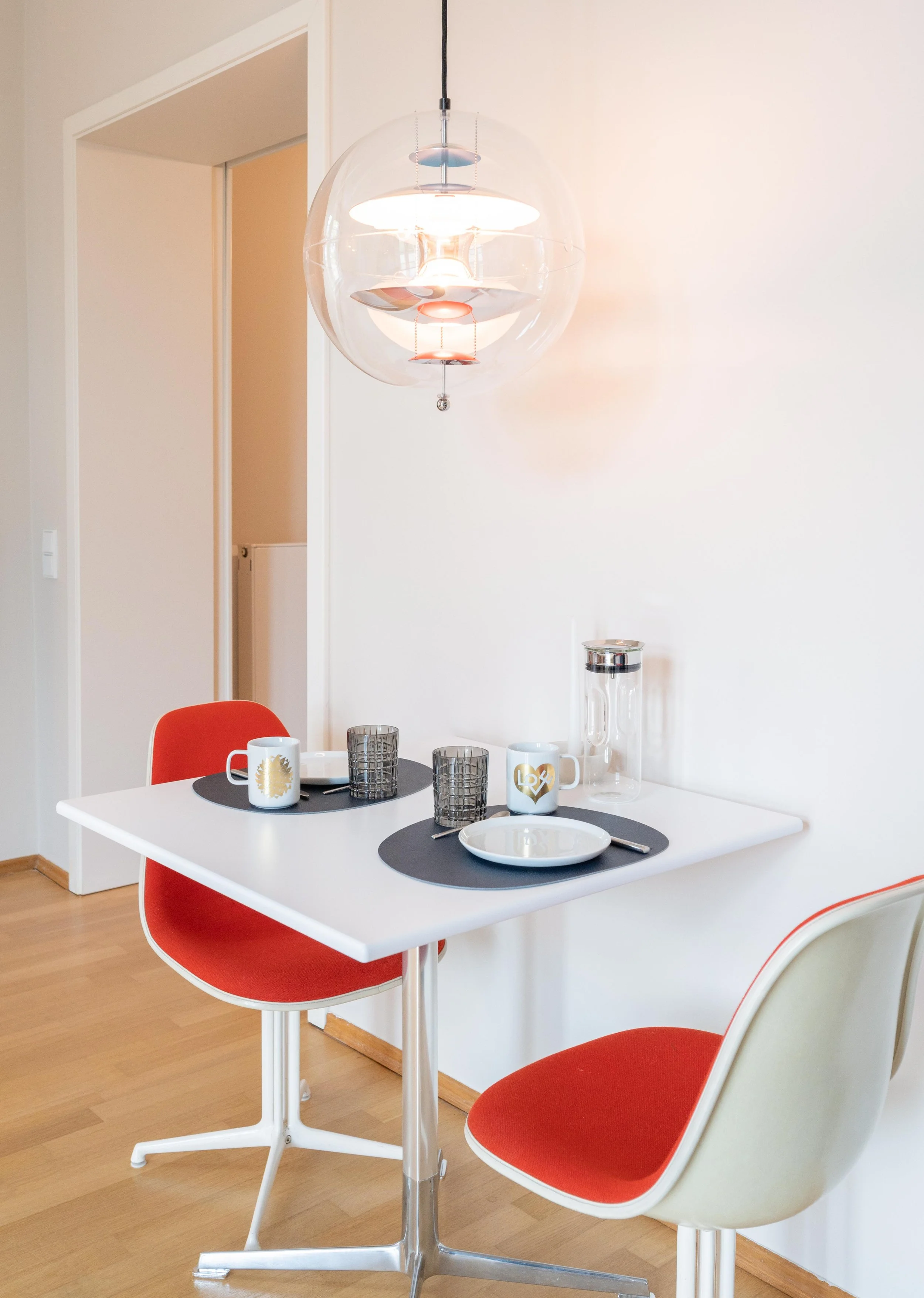 Esszimmer mit weißem Tisch, zwei roten Stühlen, schwarzen Tischsets, weißen Tassen mit goldenen und silbernen Designs, zwei grauen Gläsern, einem leeren Teller, einem Wasserkrug mit Deckel, und einer modernen Deckenleuchte.