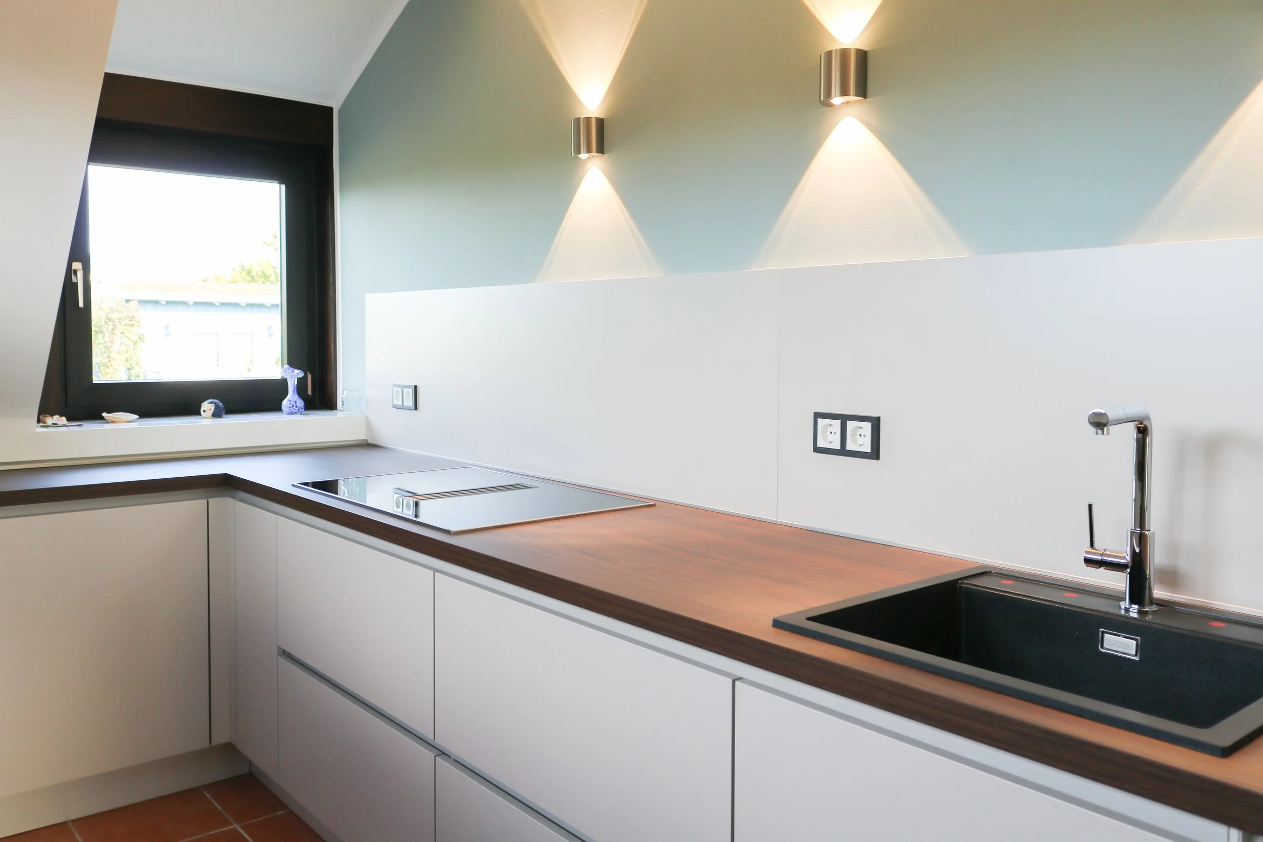 Moderne Küchenzeile mit Holzarbeitsplatte, schwarzer Spüle, Wasserhahn, Stromanschlüssen, Fenster mit Blick nach draußen, an der Wand zwei wandlampen mit Lichtprojektion nach oben und unten.