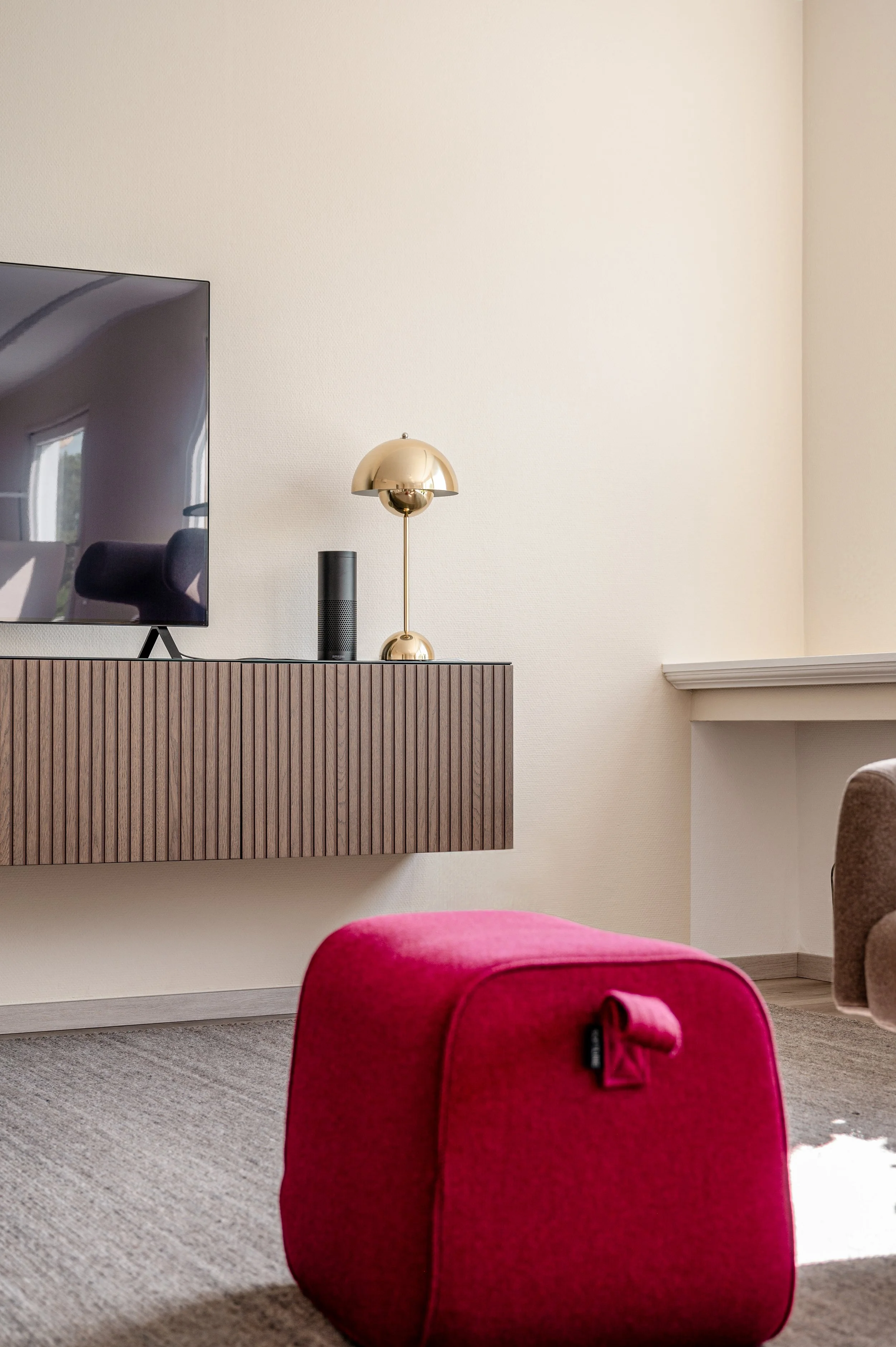 Ein roter Sitzhocker steht auf einem Teppich vor einem Fernseher, der auf einem modernen Holzschrank steht. Auf dem Schrank befindet sich eine goldene Tischlampe und ein schwarzer Lautsprecher.