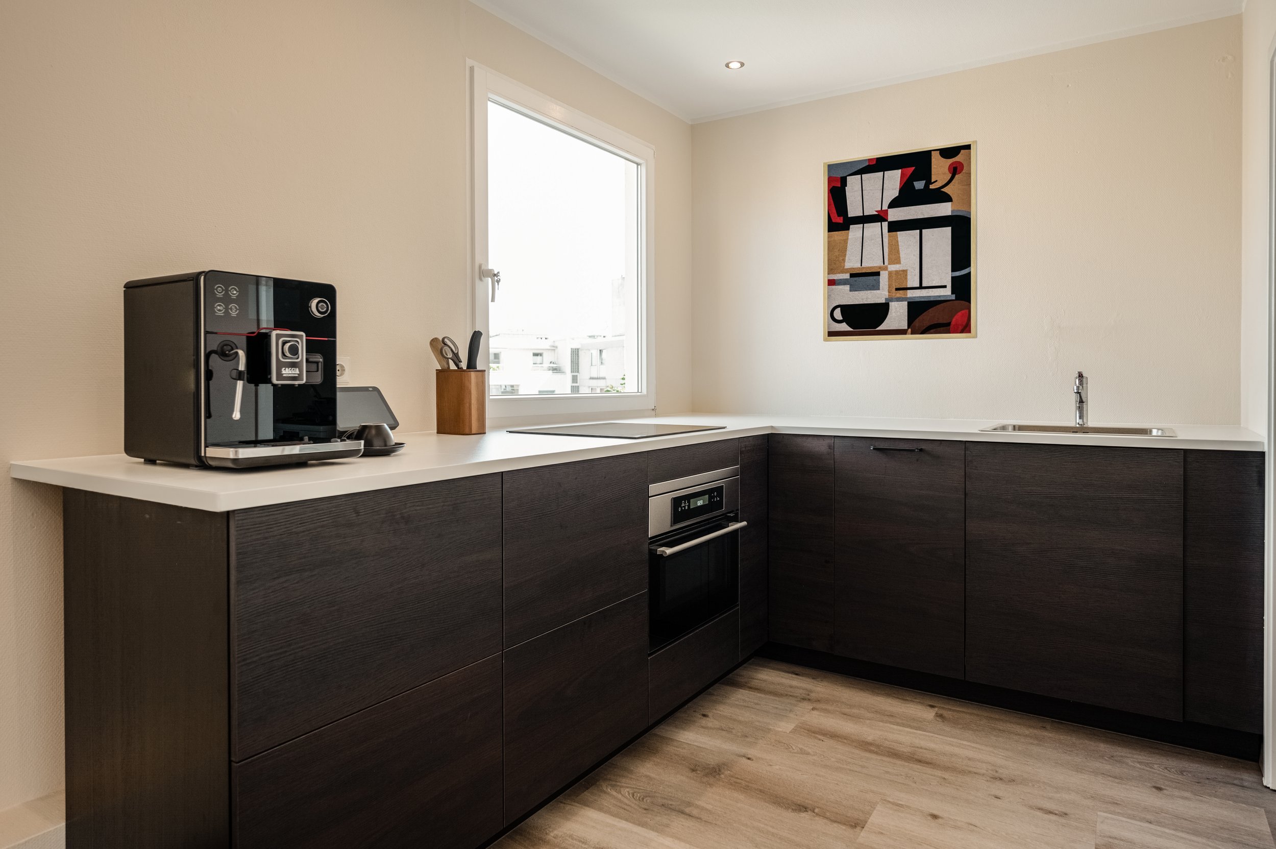 Küche mit dunklen Schränken, weißen Arbeitsflächen, Kaffeemaschine, Messerblock, Wandbild, Fenster und Spüle.