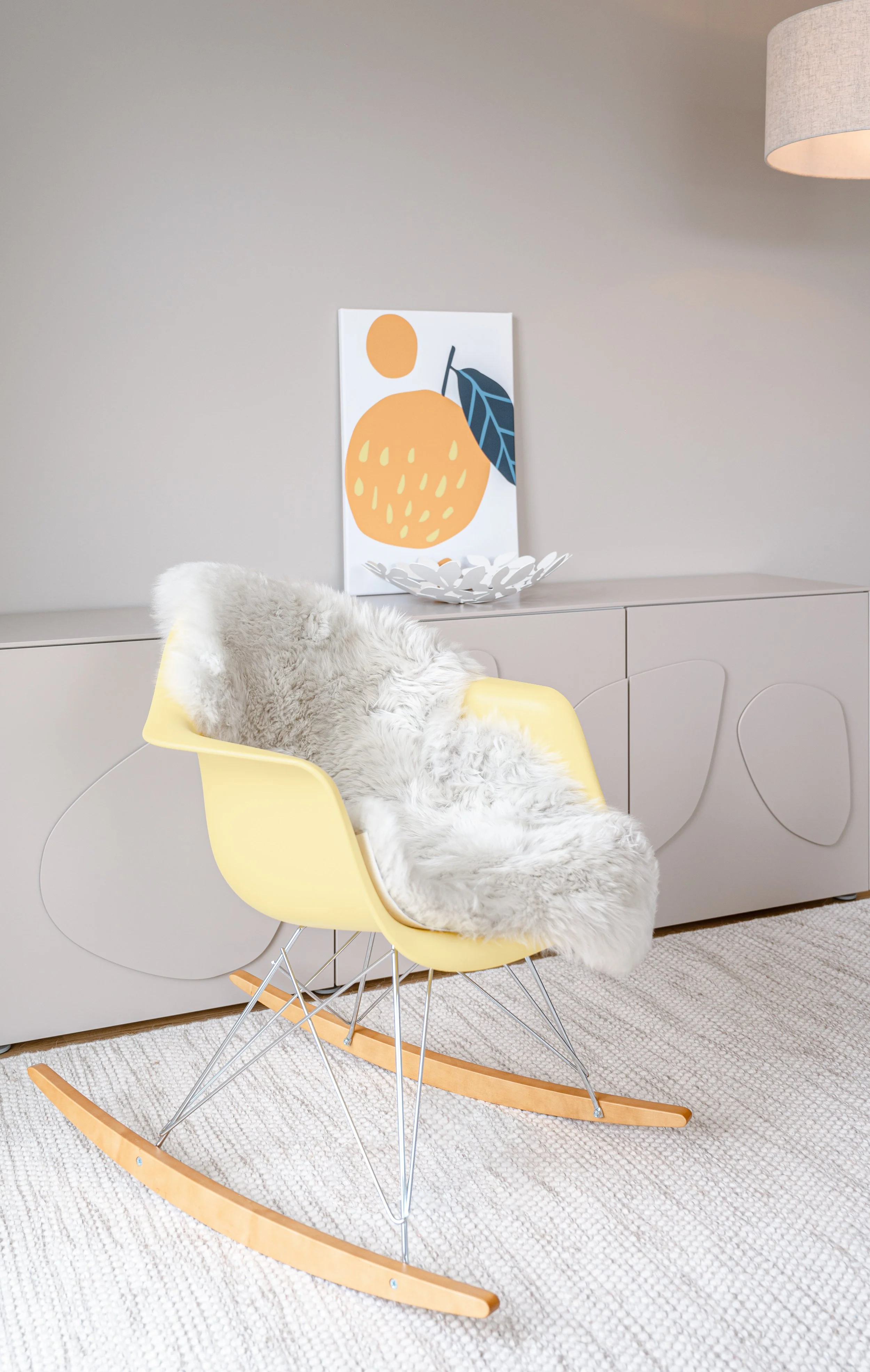 Moderner Raum mit grauer Kommode, gelbem Schaukelstuhl mit Fell, minimalistischer Wandkunst mit Orange und Blatt, grauem Lampenschirm.