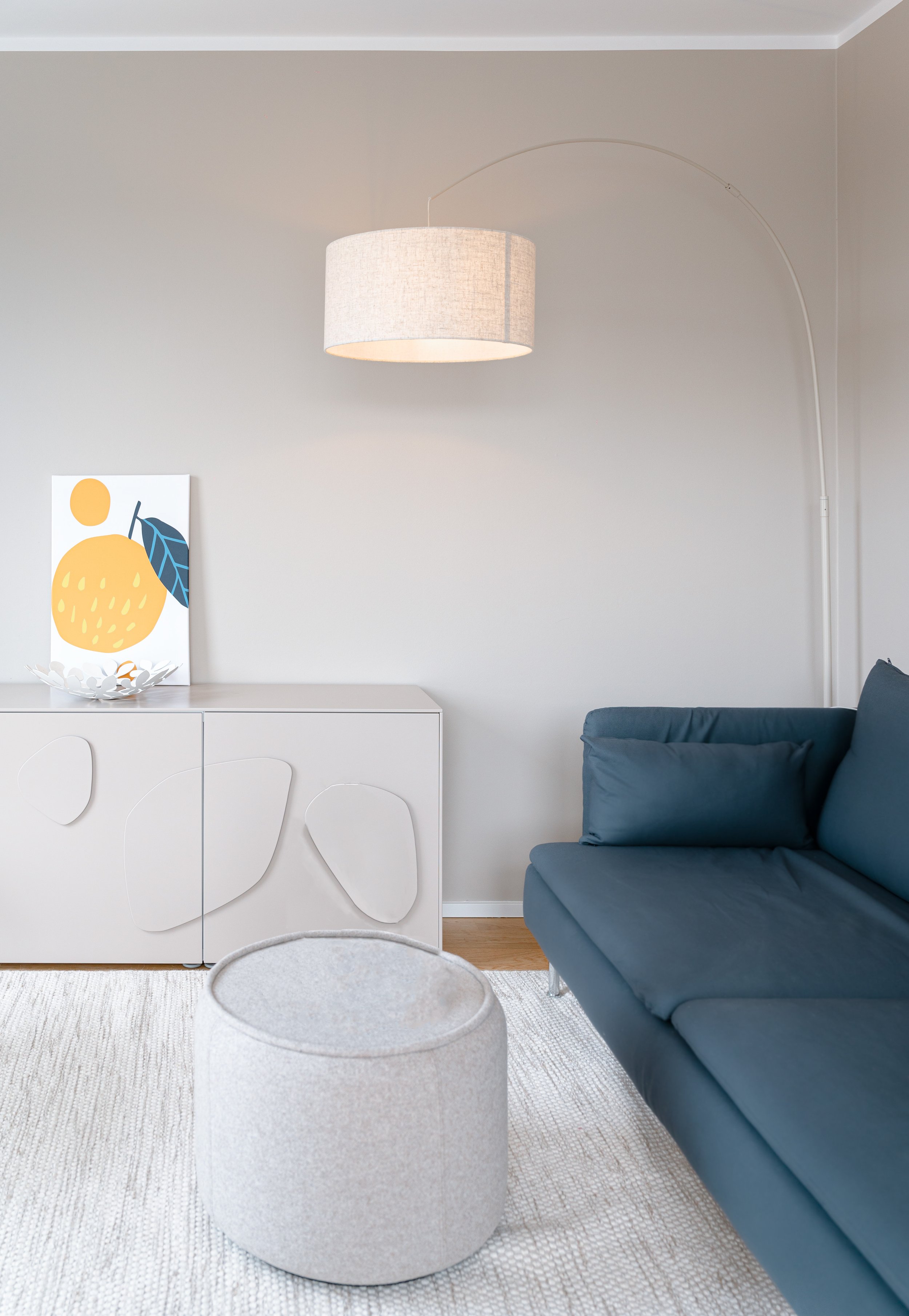 Wohnzimmer mit blauer Couch, weißen Schrank, abstraktem Kunstwerk und Stehlampe in neutralen Tönen.