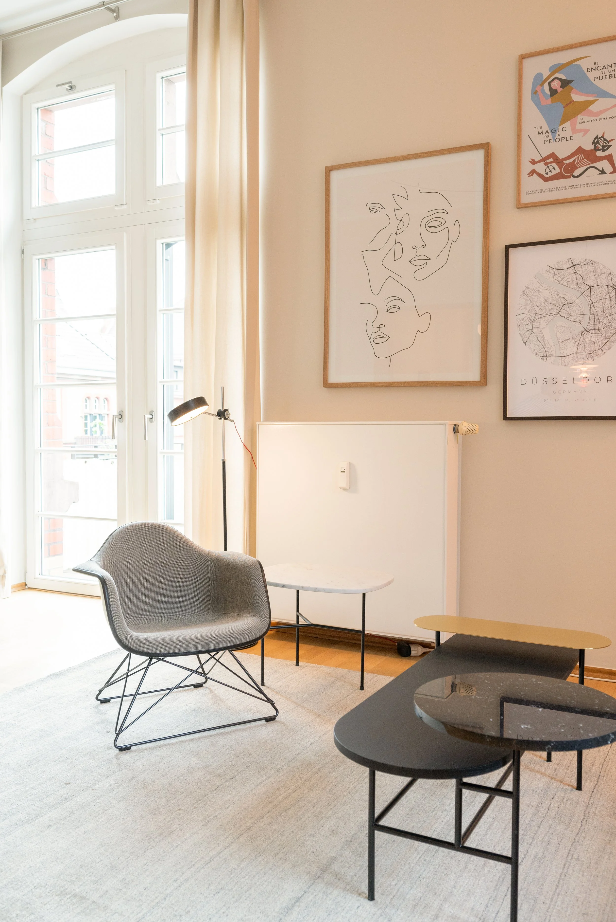 Ein modernes Wohnzimmer mit großen Fenstern, einem grauen Sessel, einem Beistelltisch und mehreren Wandbildern, darunter eine stilisierte Porträtlinie, eine Karte von Düsseldorf und ein farbiges Kunstwerk.