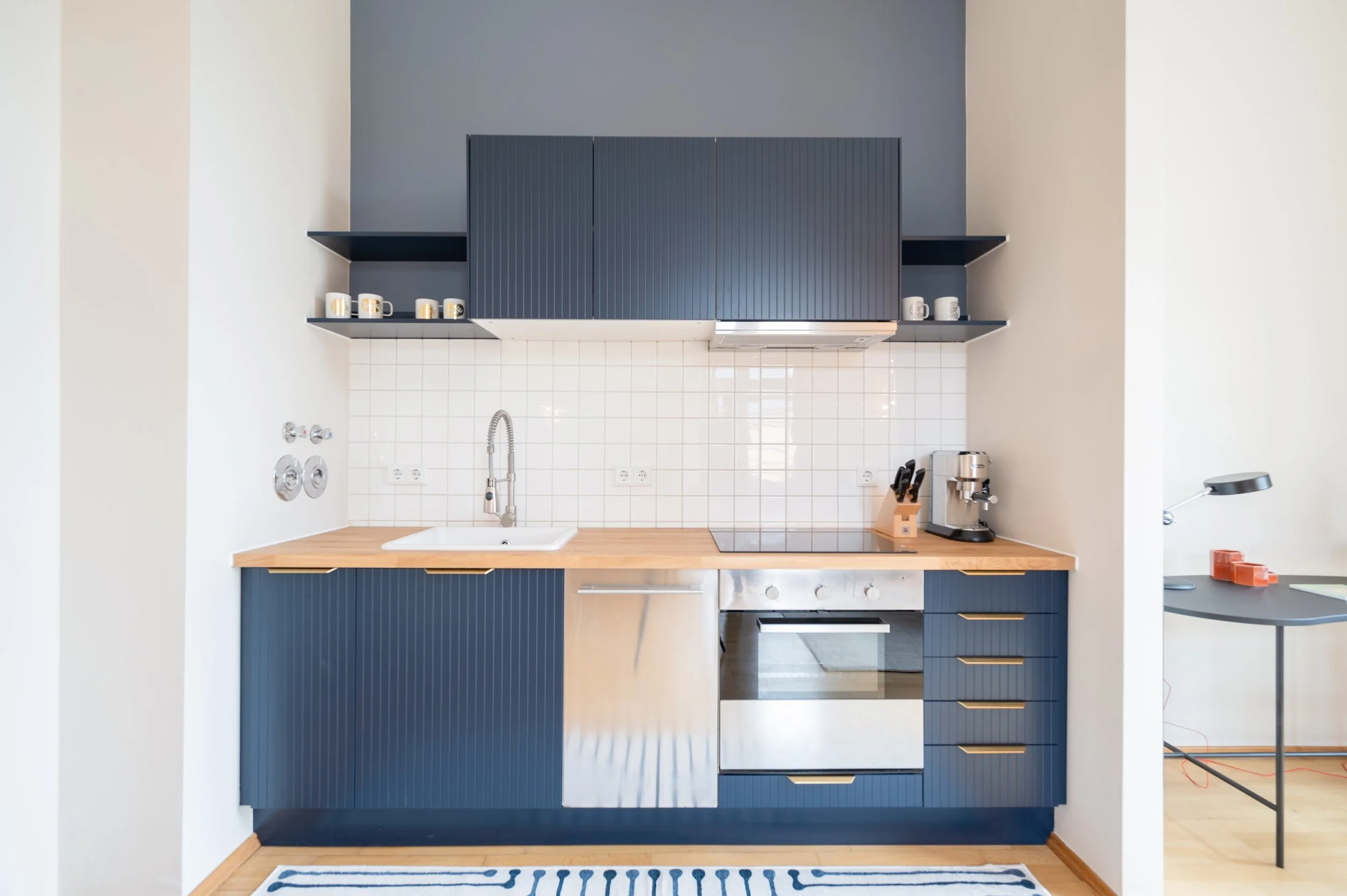 Moderne Küchenzeile mit blauen Schränken, Holzarbeitsplatte, Herd, Spüle, Kaffeemaschine und offenen Regalen mit Tassen