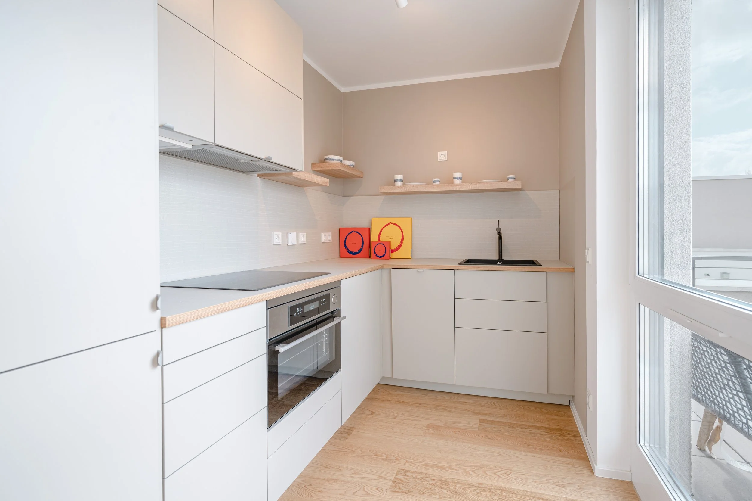 Moderne, minimalistische Küche mit weißen Schränken, Holzarbeitsplatten, einem schwarzen Spülbecken, offenen Holzregalen und einem großen Fenster, das viel Tageslicht lässt.