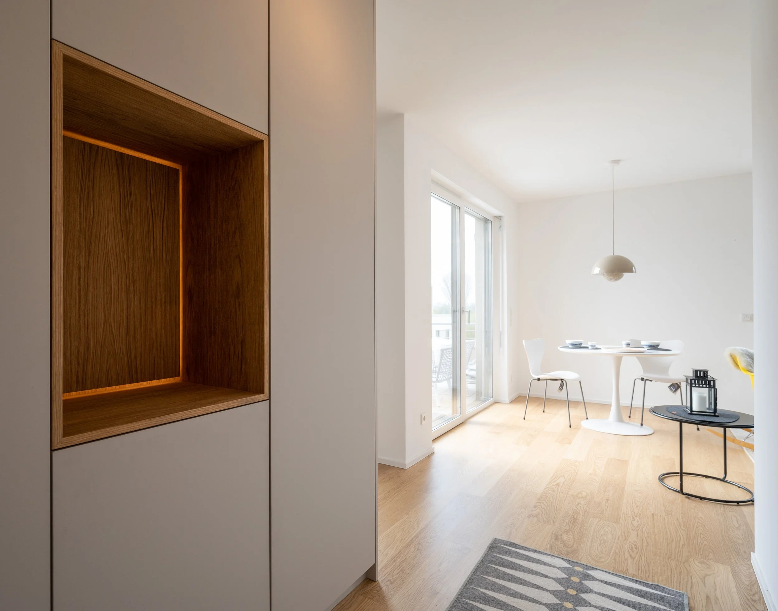 Modernes Wohnzimmer mit hellem Holzfußboden, einem weißen Esstisch mit weißen Stühlen, einem kleinen runden Beistelltisch mit schwarzem Rahmen, schwarzer Laterne und einer modernen weißen Pendelleuchte